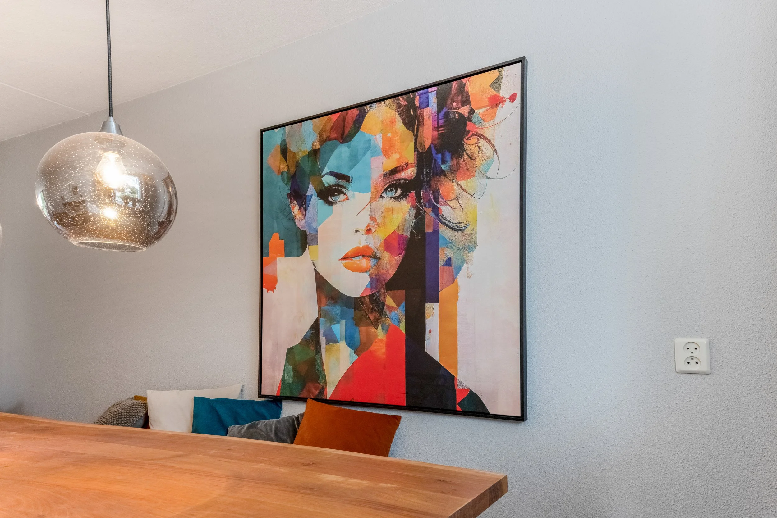 Een kleurrijk schilderij van een vrouw met kort haar op een witte muur, naast een houten tafel met kussens.