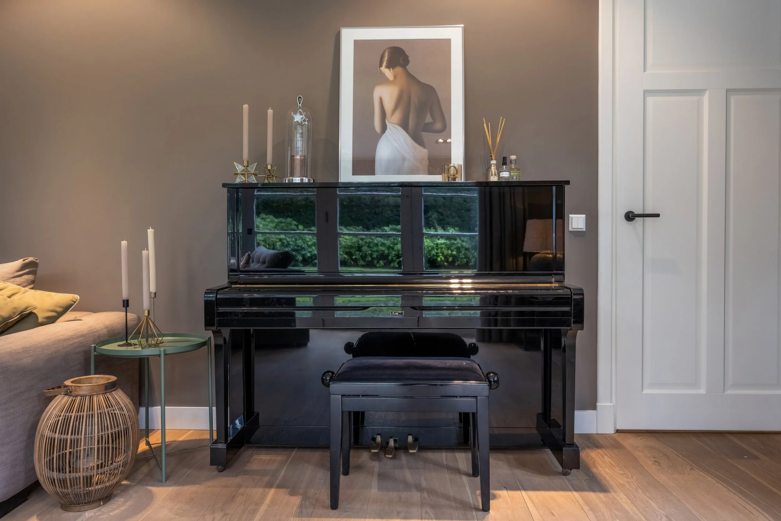 Een zwarte piano met een bijpassende zitbank, geplaatst voor een grijze muur met een kunstwerk van een vrouw met haar rug naar de camera. Boven de piano staan decoratieve items, waaronder kaarsen, een glazen fles en geurverspreiders. Aan de linkerkan