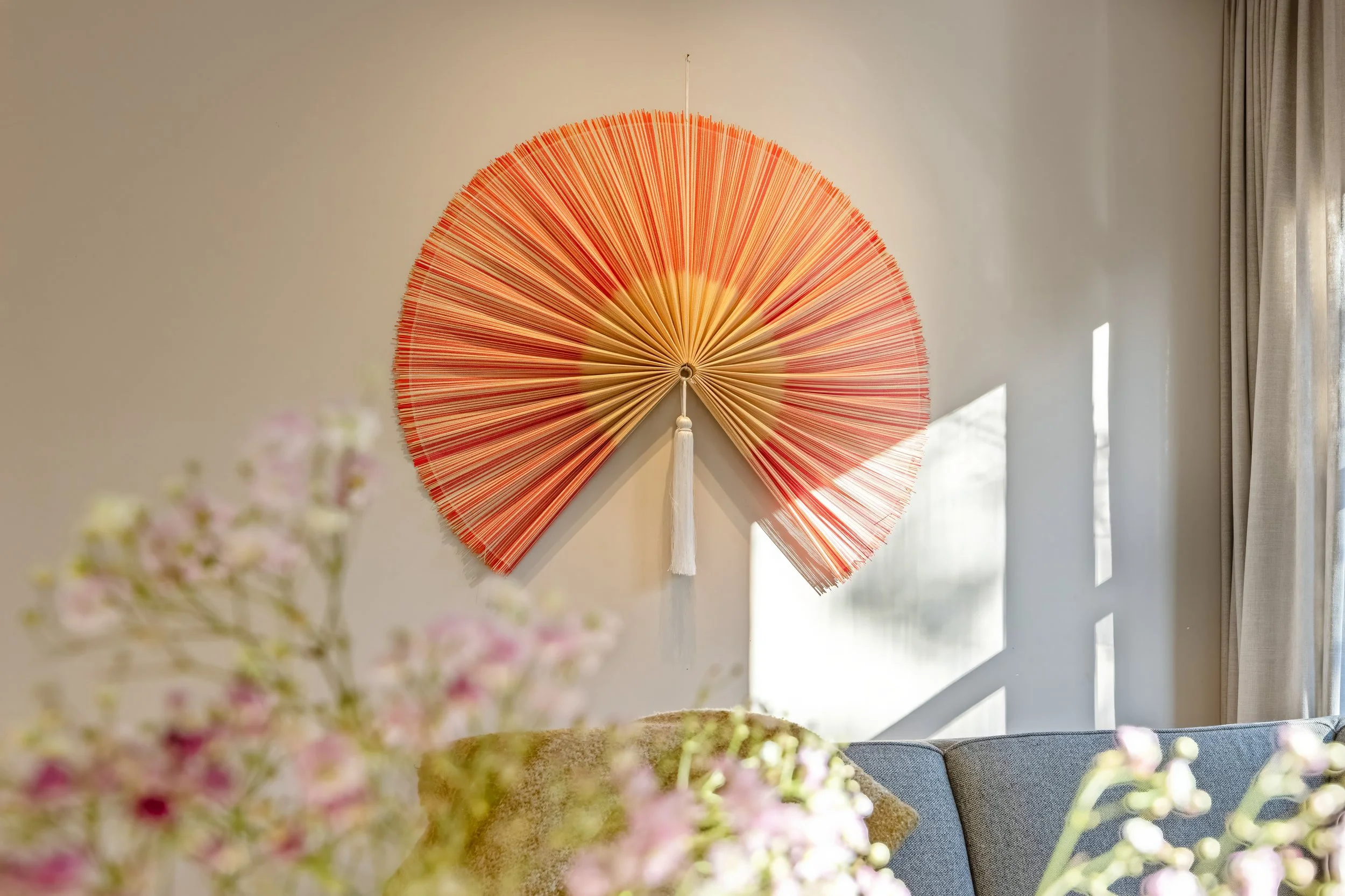 Decoratieve ronde wandlamp met rood en oranje lijnen, hangend op een witte muur naast een raam met gordijnen, met bloemen op de voorgrond.
