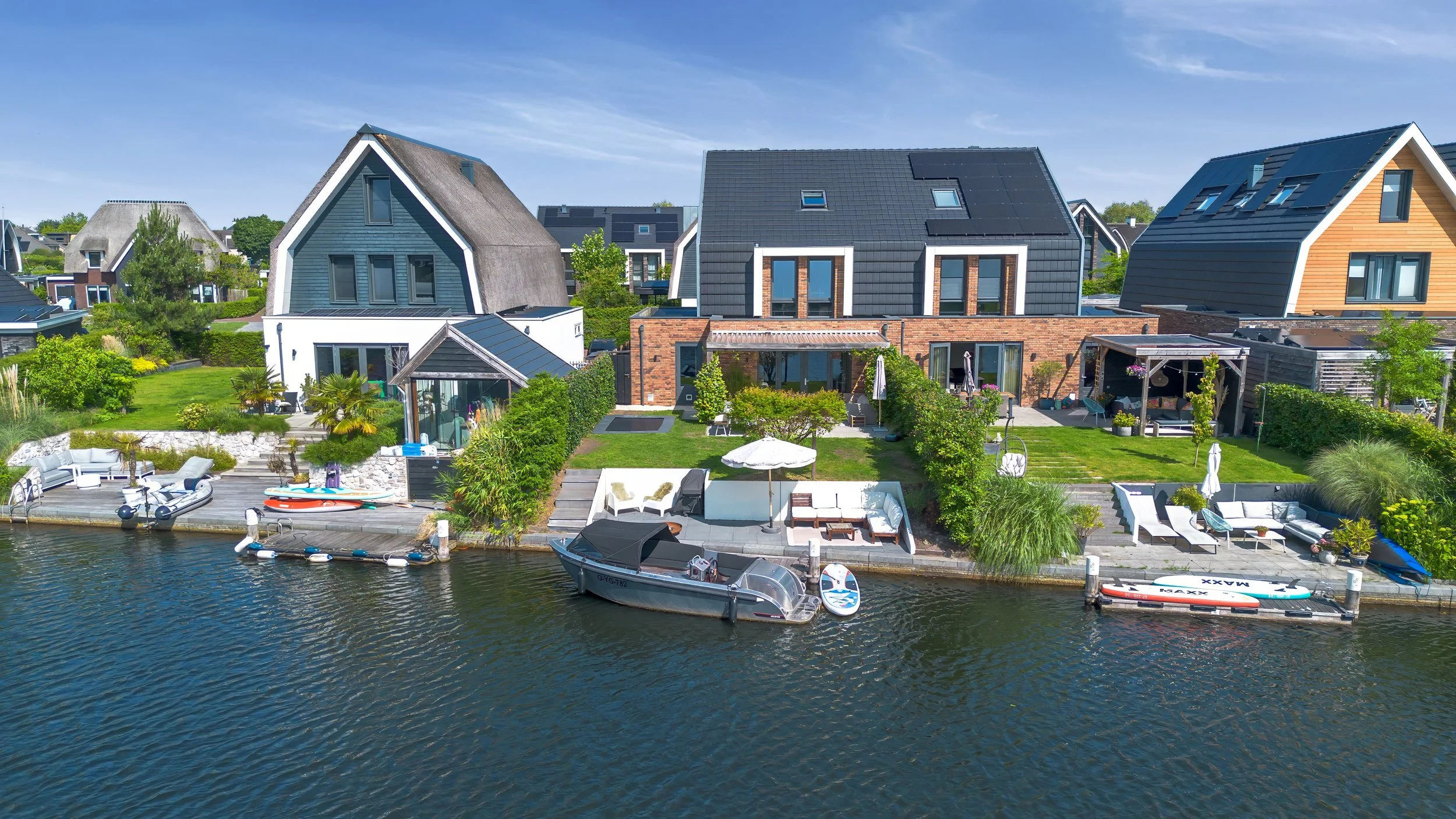Gezicht op moderne huis aan het water met tuin, ligplaatsen voor boten, ligbedden en terras met parasols en lounge meubels.