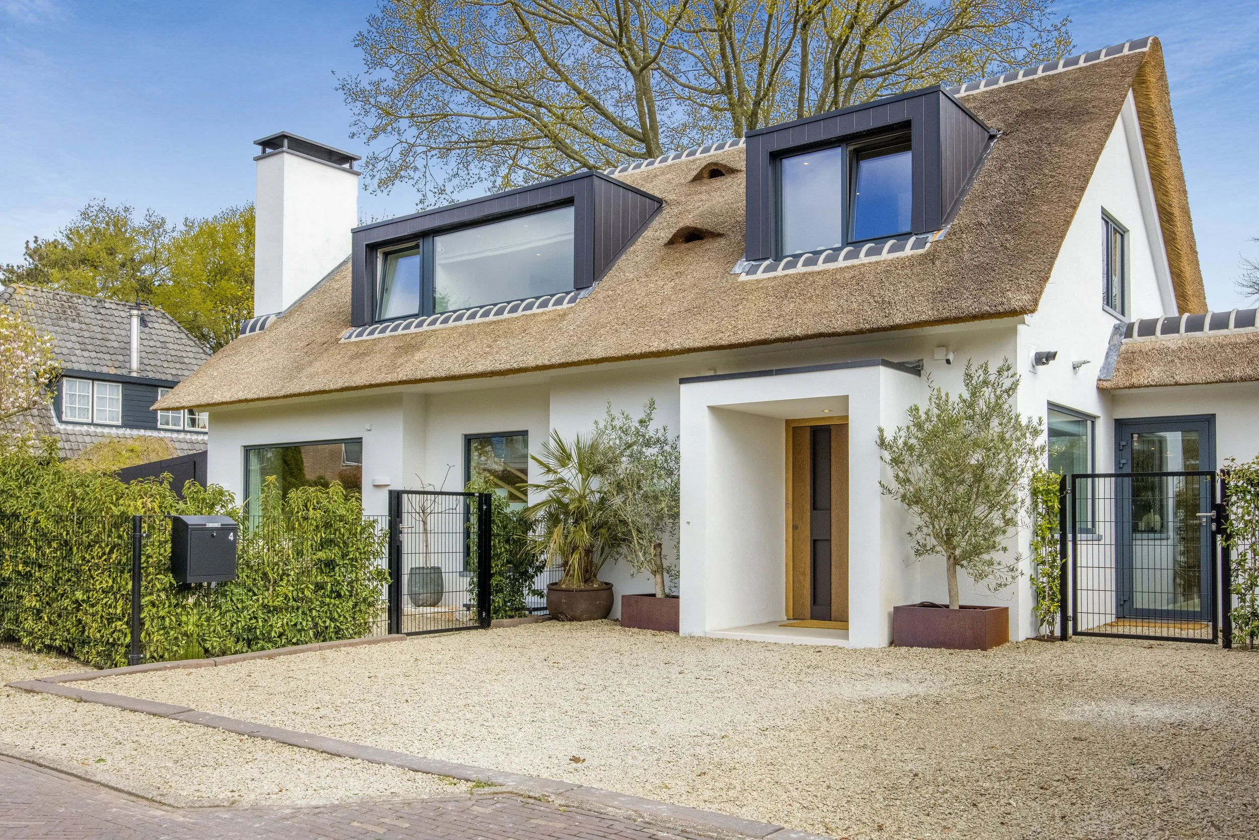 Moderne witte woning met een rieten dak en grote ramen, omgeven door een tuin met planten en een grindpad.
