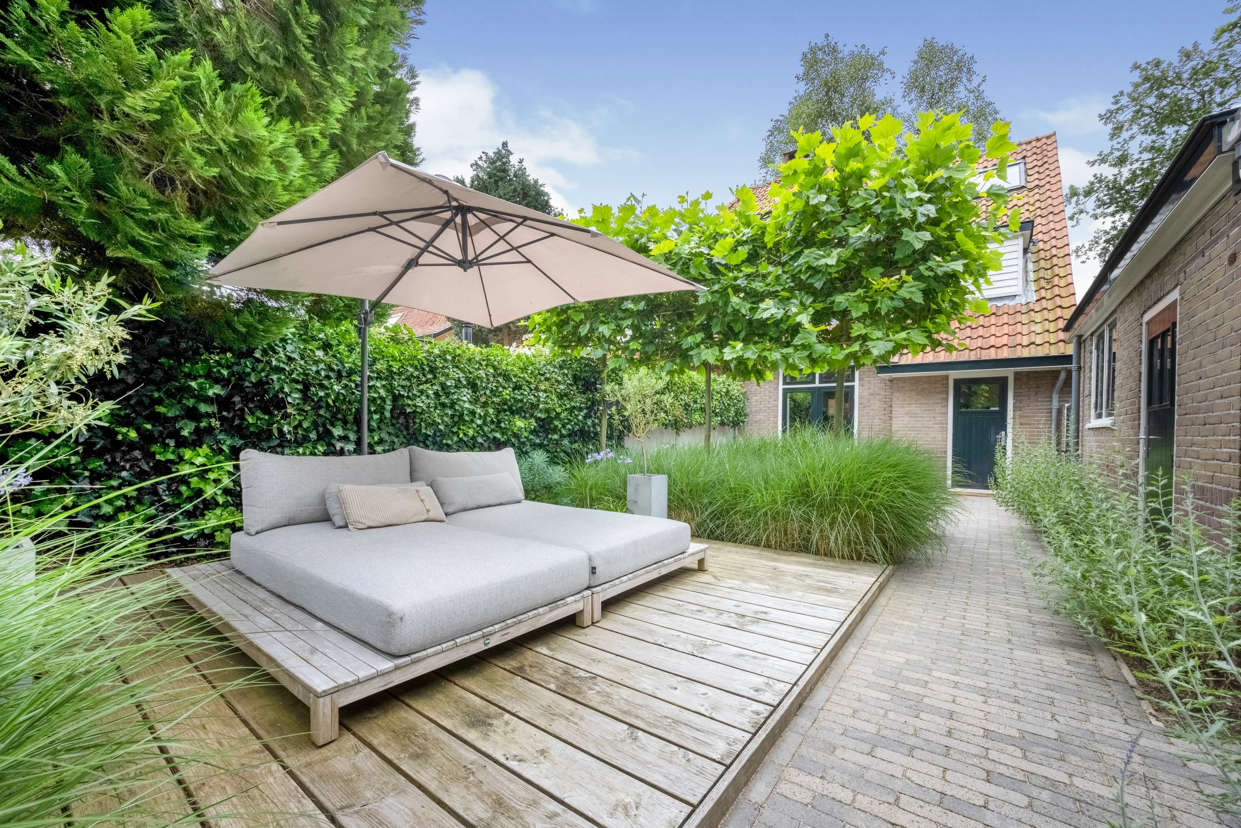Een zithoek met een loungebank onder een grote parasol op een houten terras, omgeven door groene planten en bomen, met een bakstenen huis op de achtergrond.