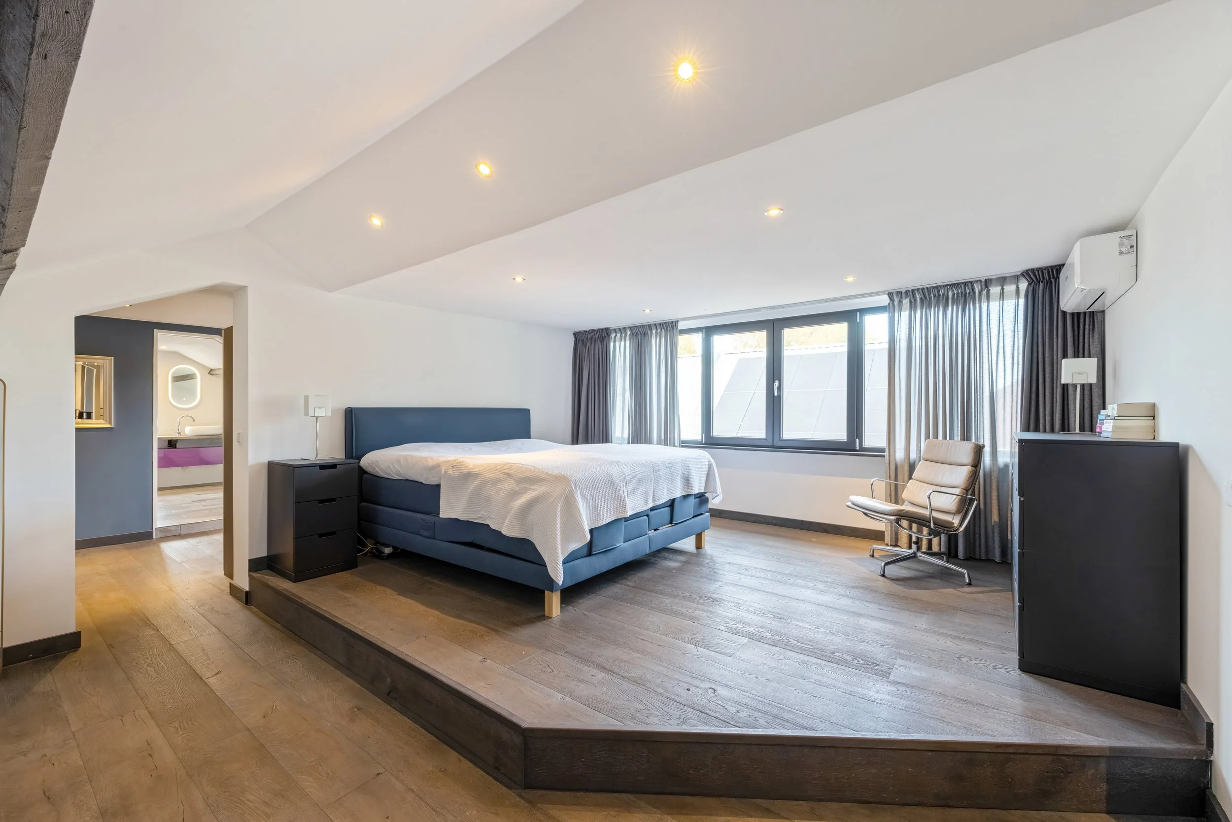 Ruimtelijke slaapkamer met groot bed, grote ramen met gordijnen, beige stoel, zwarte kast, airconditioning, houten vloer, plafondverlichting.