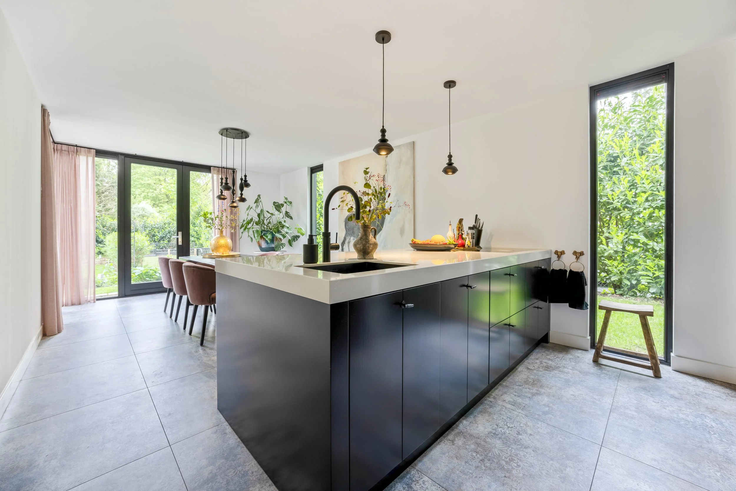 Moderne keuken met zwart keukenblok, witte werkblad, centraal eiland, planten bij groot raam, eetgedeelte met roze stoelen, grote ramen en tuin te zien.