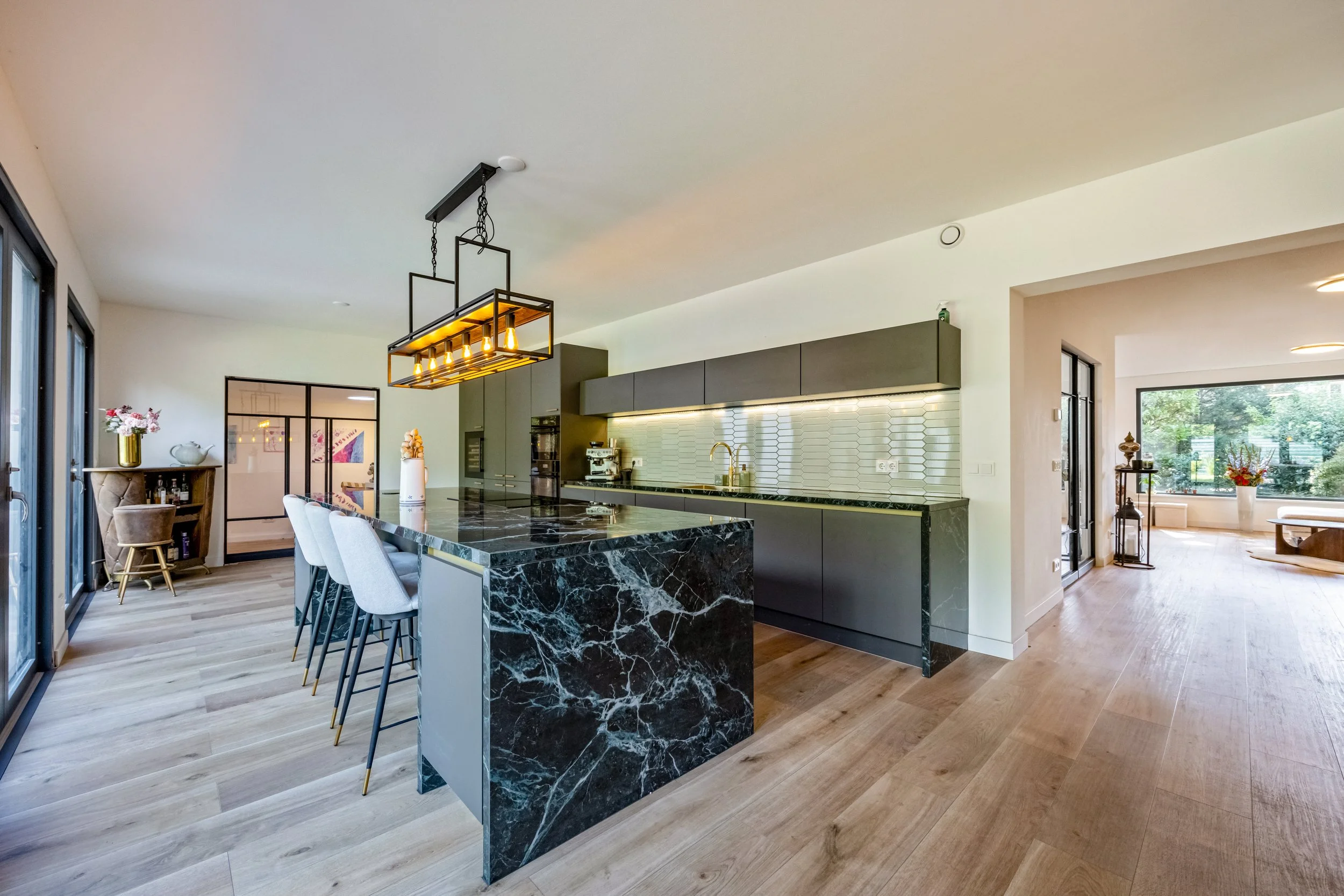 Modern keuken met zwart marmeren eiland, grijze kasten, en een lichte houten vloer, grote ramen voor natuurlijk licht.