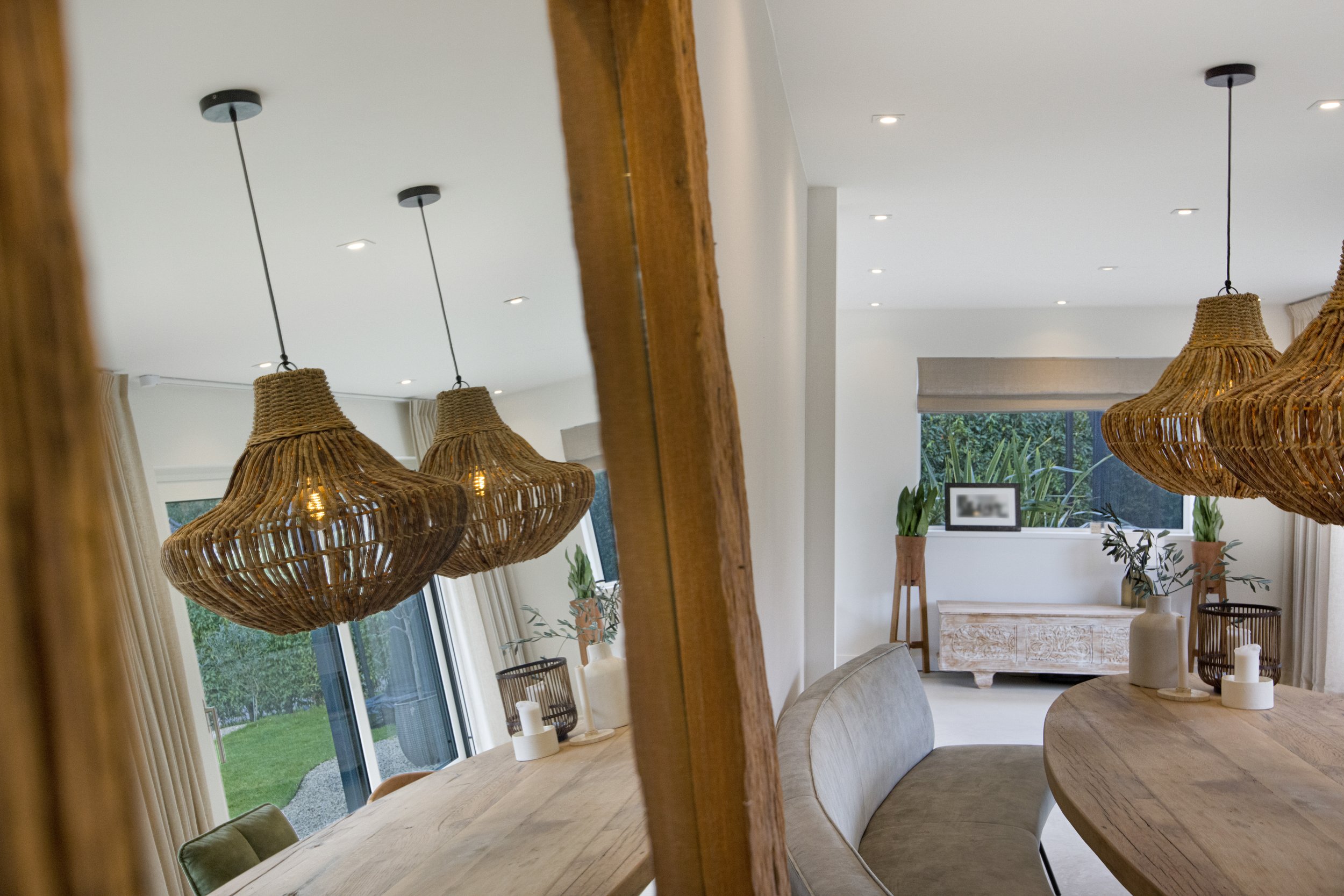 De afbeelding toont een interieur van een moderne woonruimte met een grote houten eettafel, omringd door een bank en stoelen, met opvallende rieten hanglampen erboven. Achter in de ruimte zijn grote ramen met uitzicht op een tuin, en binnen staan pla