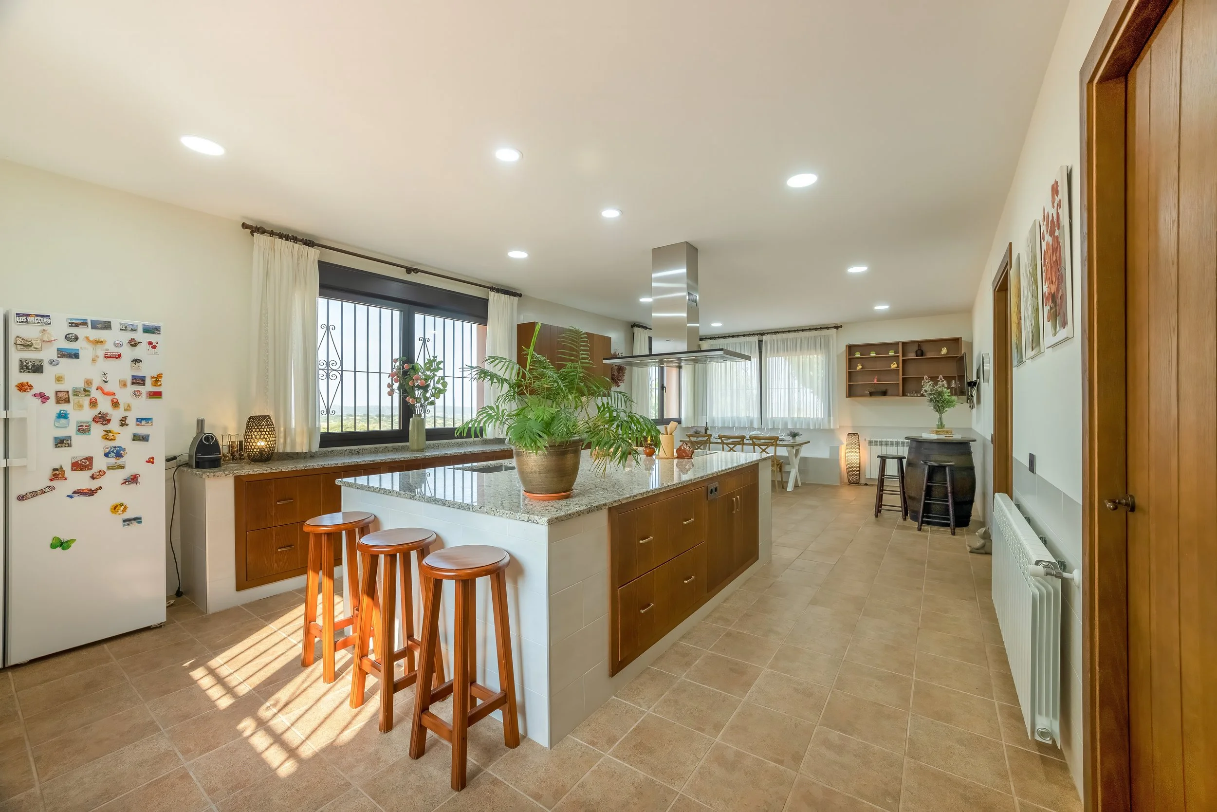 Ruime keuken met centrale eiland, houten stoelen, koelkast met magneetjes, grote ramen met uitzicht en vitrage, decoratieve planten en lampen, eettafel en wandplanken met decoratie.