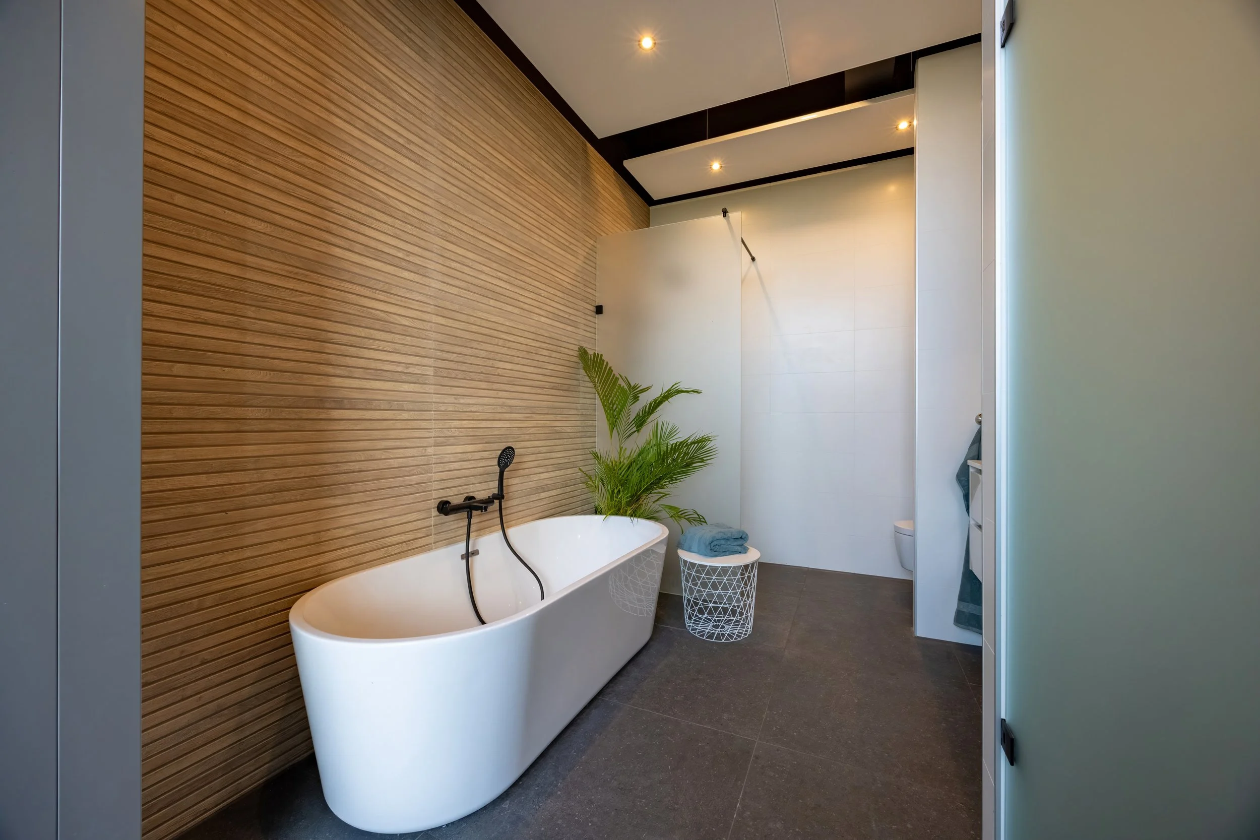 Moderne badkamer met vrijstaand bad, palmplant, wasmand met handdoek, en inloopdouche op de achtergrond.