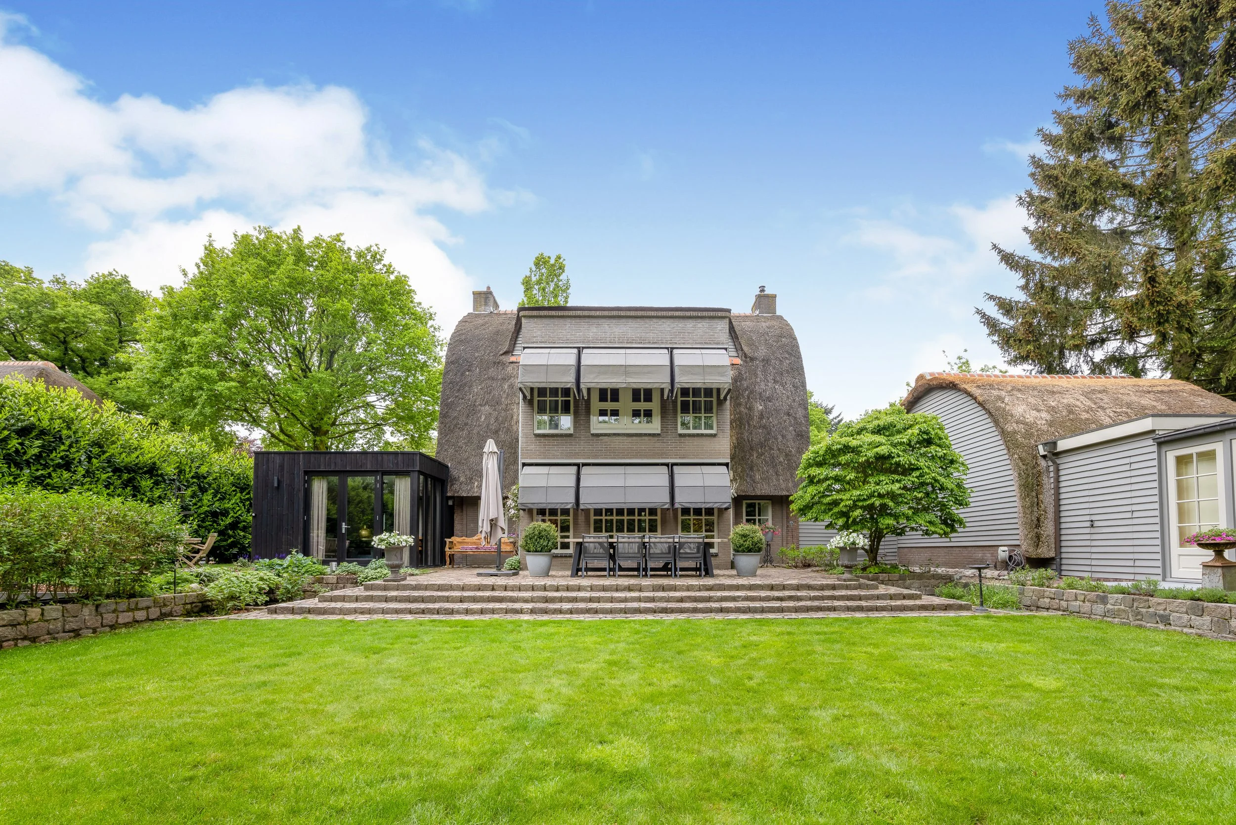 Een huis met een datsja-achtig dak en een groene tuin met een trappenhuis, tuinmeubelen, planten en bomen.