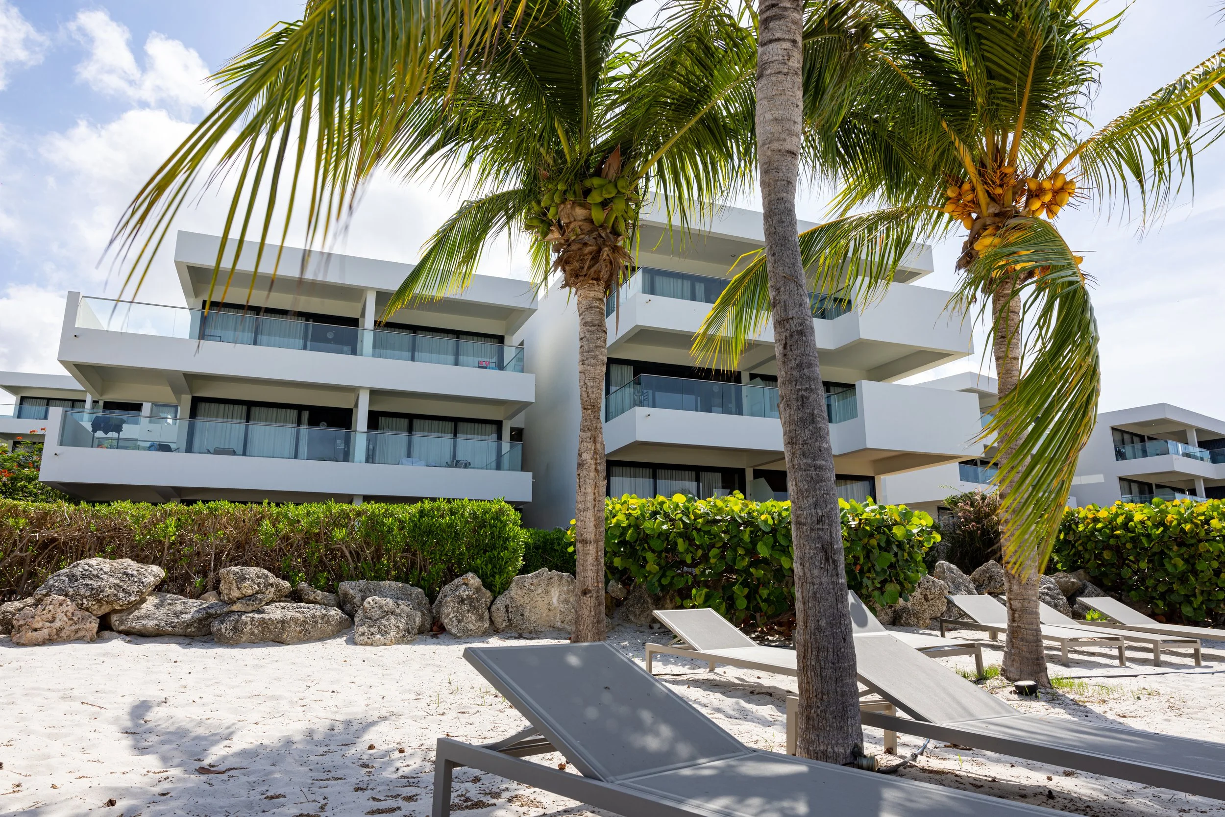 Modern witte appartementen met grote balkons, palmbomen en ligstoelen op het strand in een zonnige omgeving.