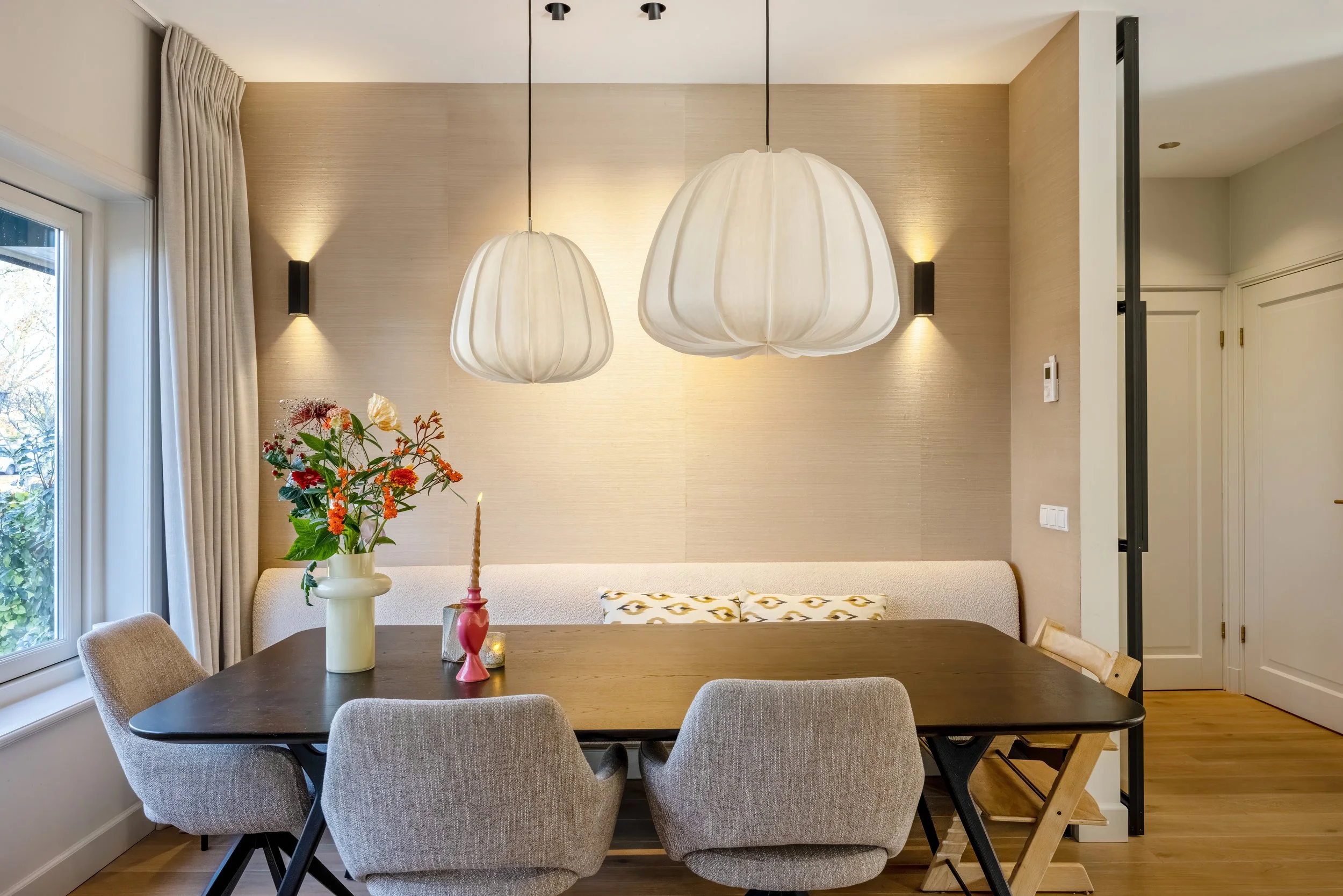 Eettafel met vazen met bloemen en een kaars, drie grote plafondlampen, beige muren en gordijnen, houten vloer, lichte muurkleuren.