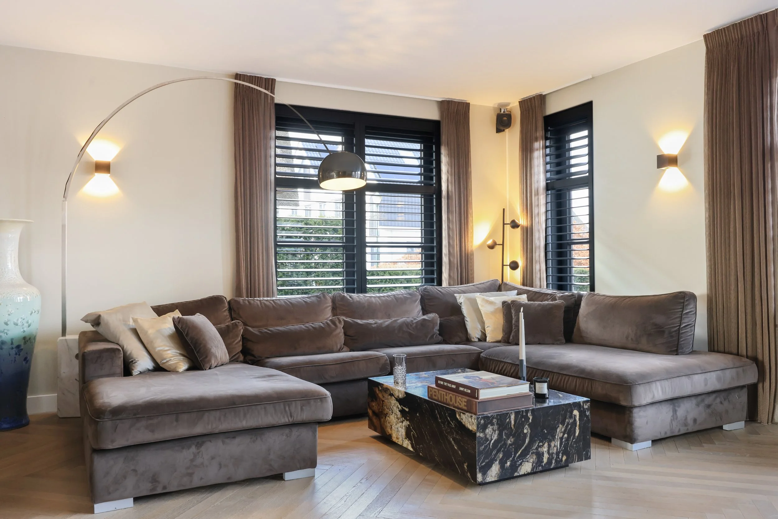 Een woonkamer met een grote grijze bank, een zwart marmeren salontafel, grote ramen met zwarte jaloezieën, beige gordijnen, een plafondlamp en muurpendelverlichting.