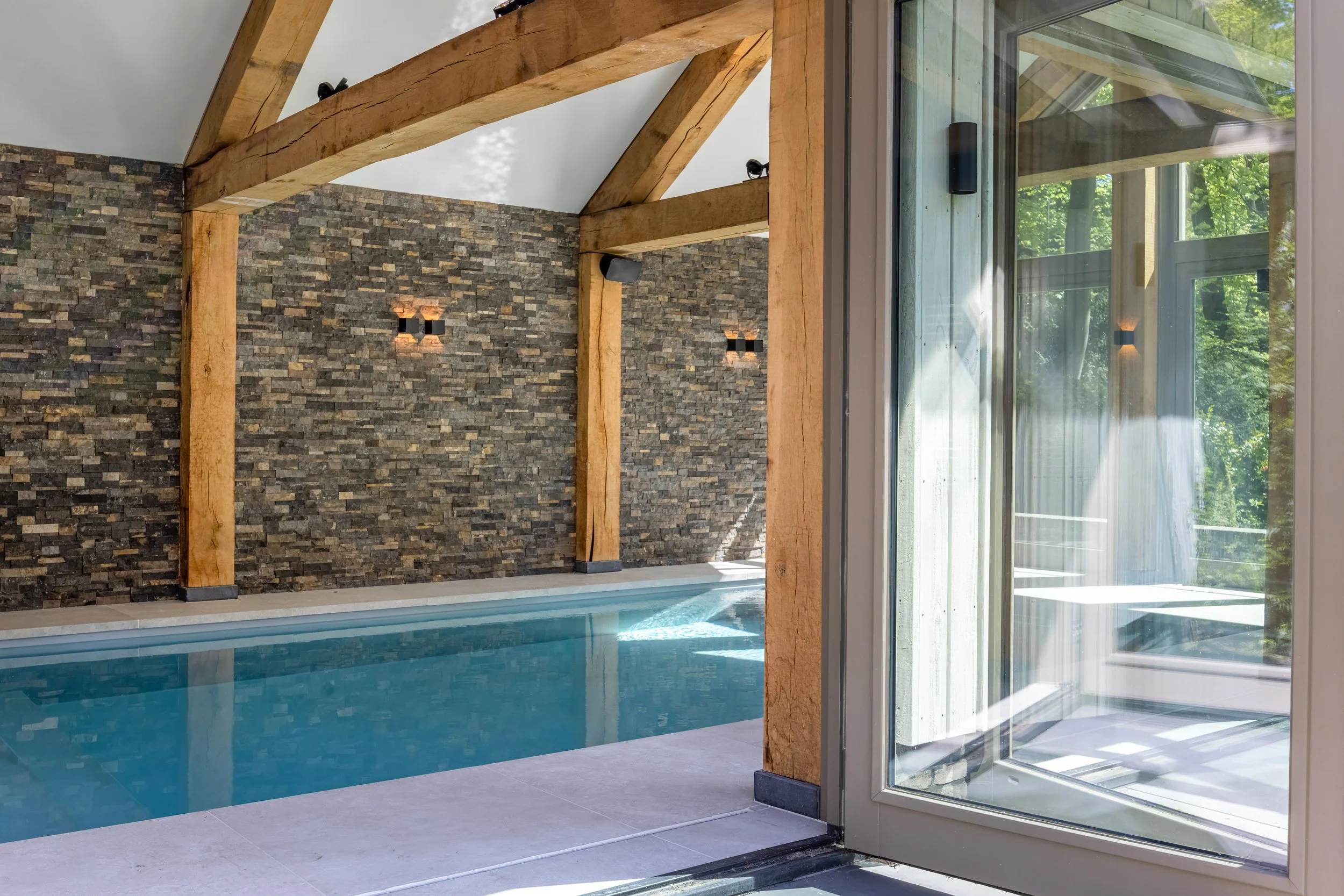 Binnenzwembad met stenen muur en houten dragende balken, grote glazen schuifdeur naar een terras met uitzicht op een groene tuin.
