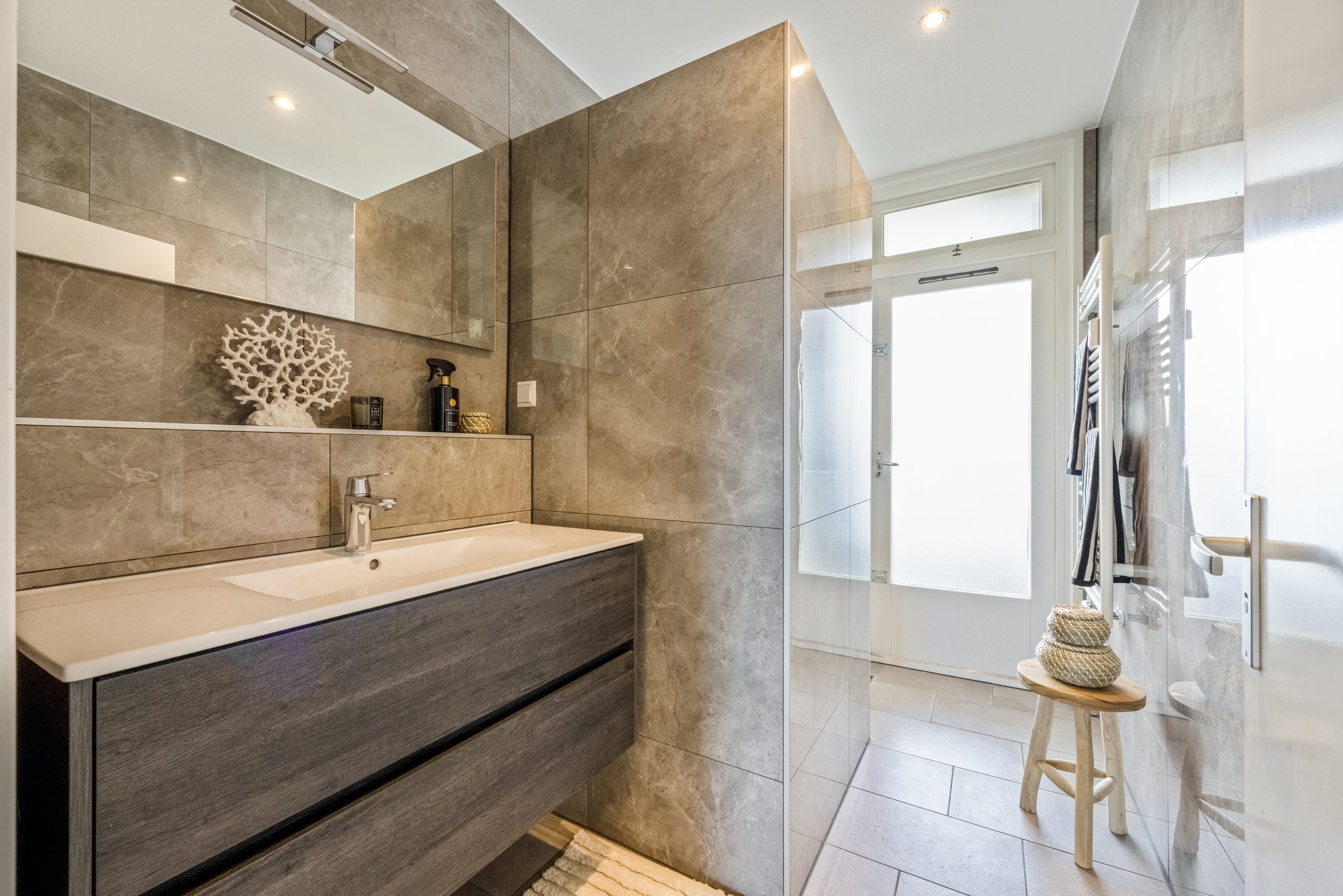 Moderne badkamer met grote spiegel, wastafel met lade, spiegelkast, badlijn en handdoekradiator, deur naar balkon, decoratieve zee-coralmantel, natuurlijke kleuren en lichte houten stoel.