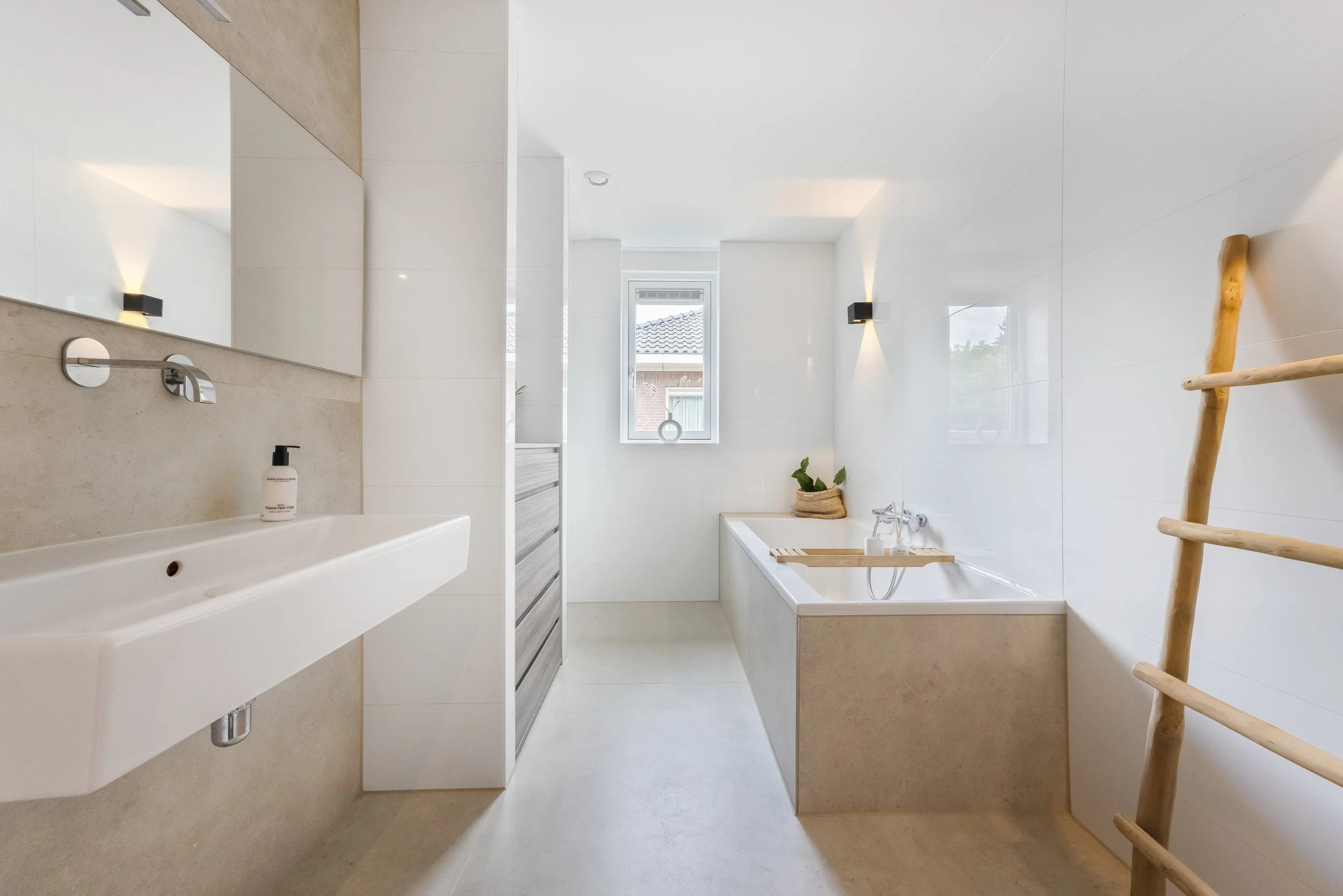 Een minimalistisch witte badkamer met een wastafel, bad, ladder en een smallere kast, verlicht door wandlampen en natuurlijk licht via een raam.