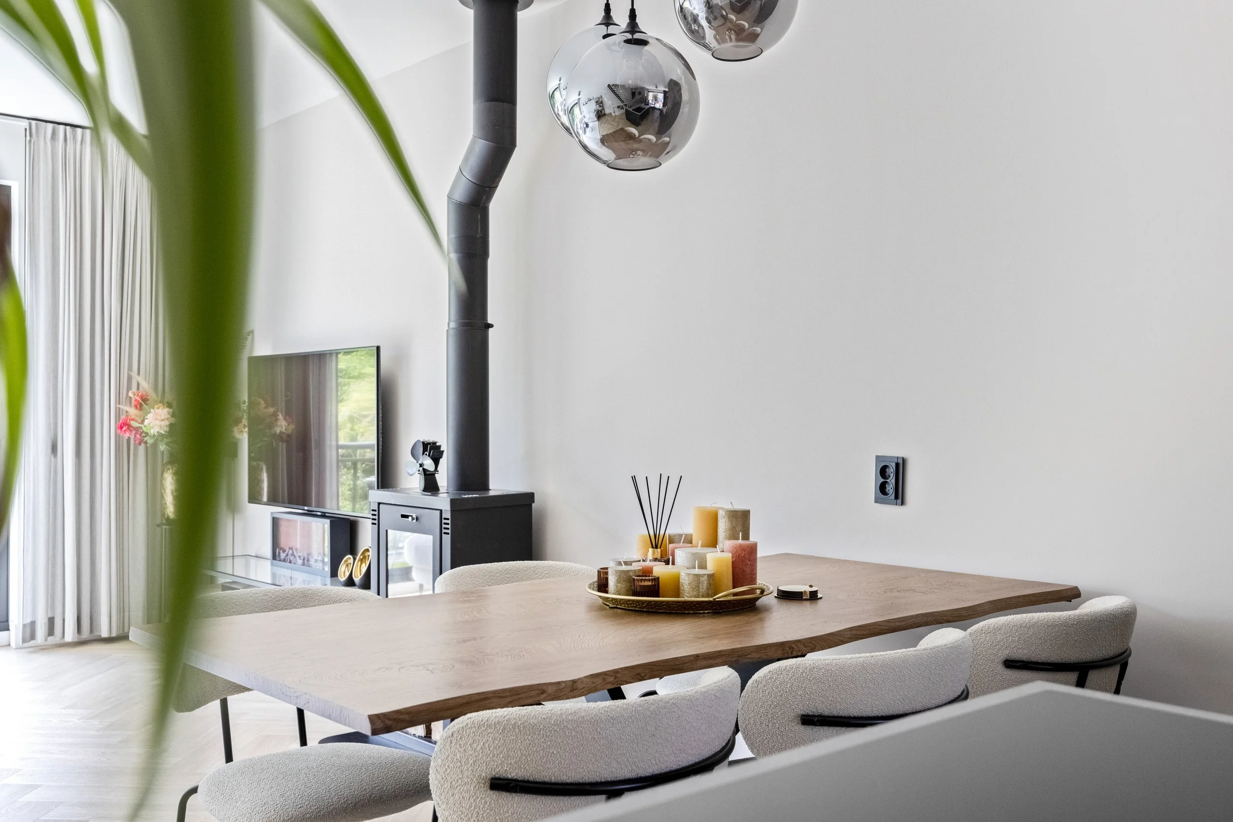 Eethoek met houten tafel, witte stoelen, kaarsen op dienblad, moderne woonkamer met tv, open haard en glazen deur met gordijnen.