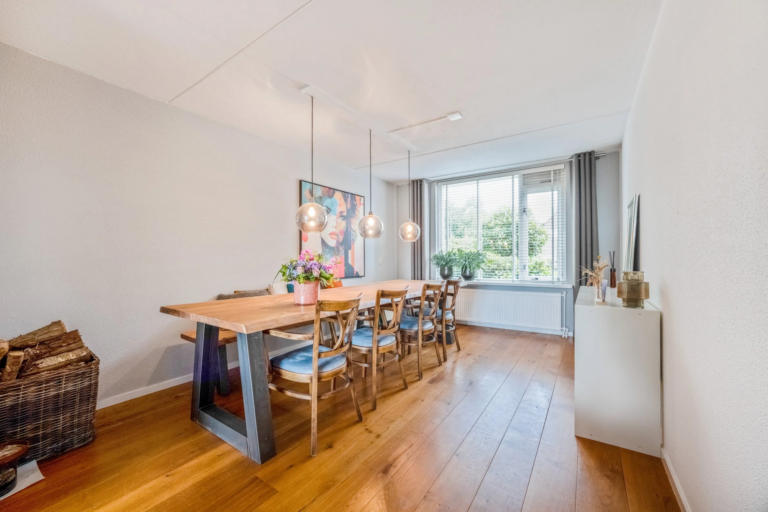 Eetkamer met houten tafel, zes houten stoelen, bloemen op tafel, grote raam met gordijnen, muurkunst, en planten op vensterbank.