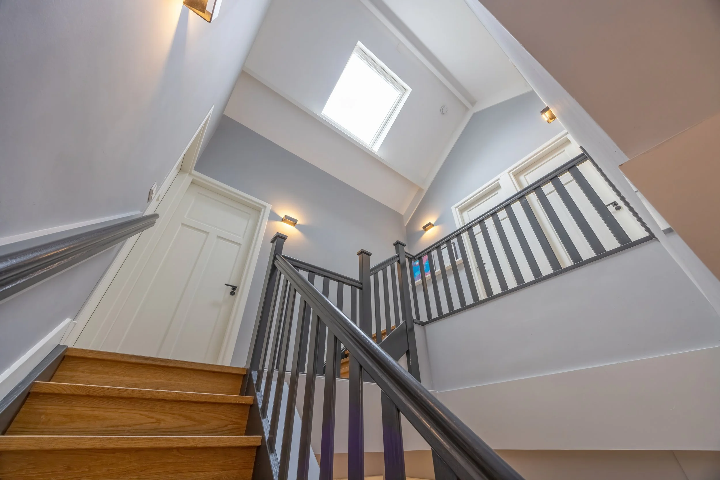 Interne trap in een modern huis met houten treden, een zwarte balustrade en een plafond met een venster dat daglicht binnenlaat.