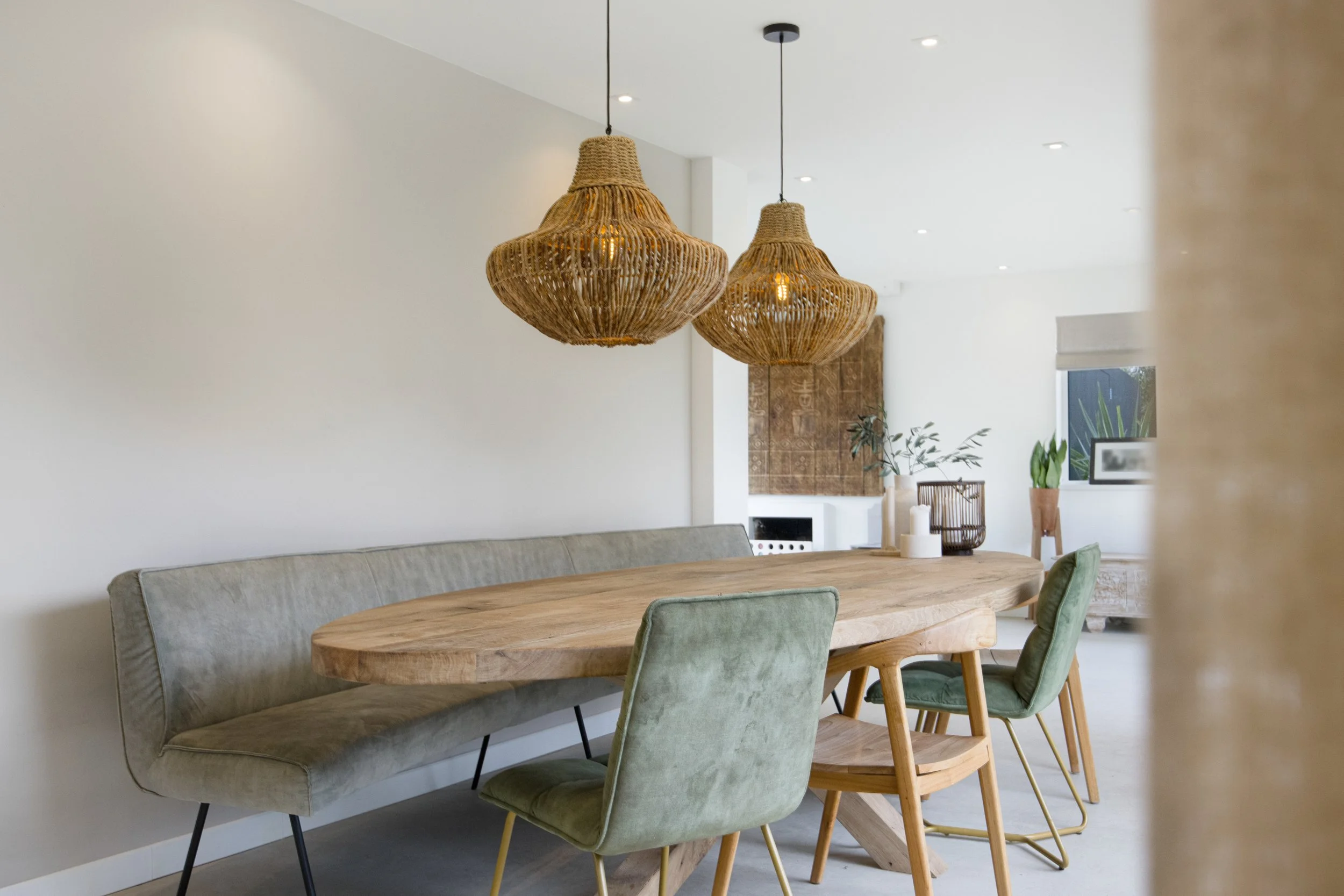 Lichte eetkamer met houten tafel, groene en houten stoelen, gebladerde hanglampen, witte muren en planten in de achtergrond.