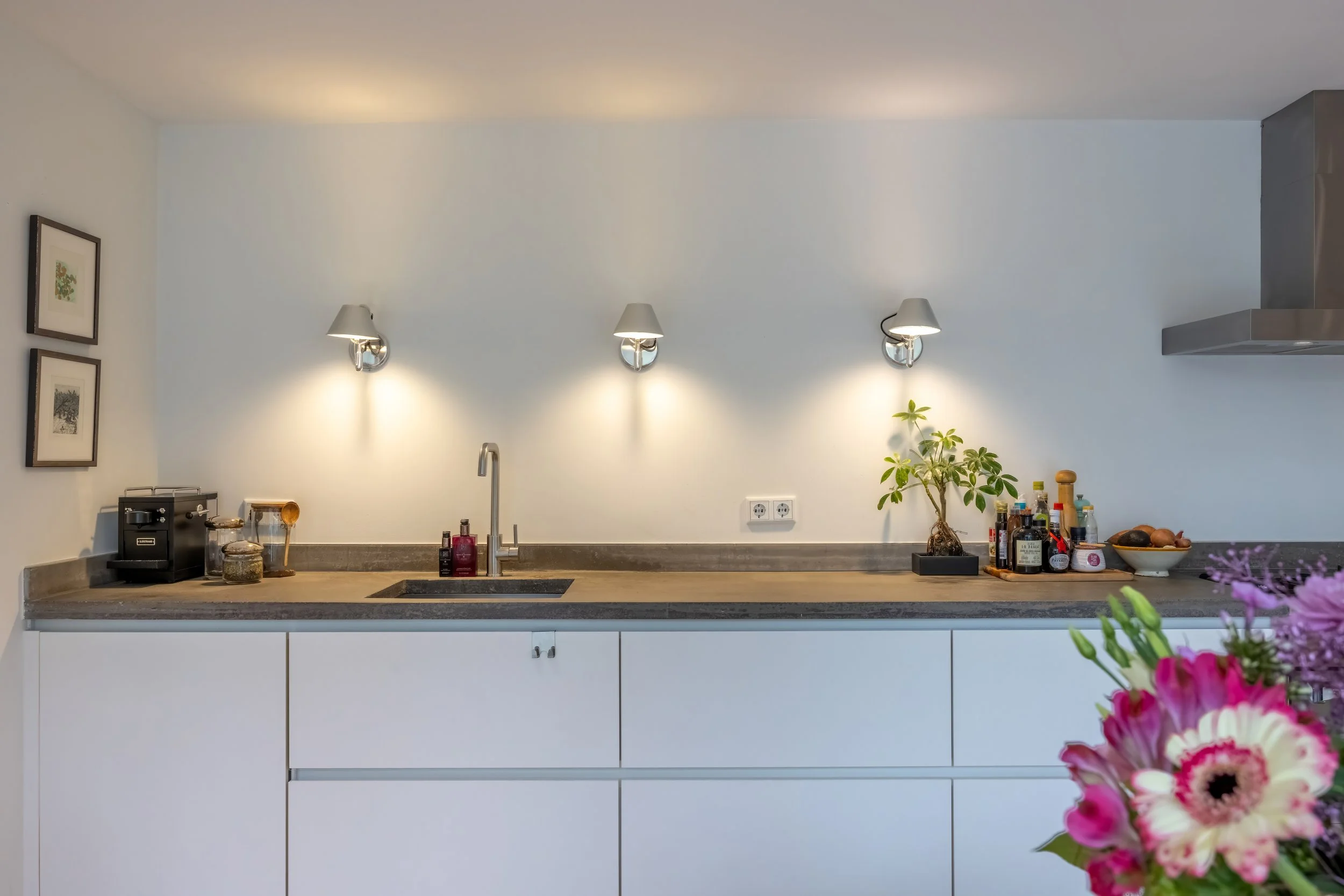Een moderne keuken met witte kasten, een grijze werkblad, een gootsteen, drie muurlichten, een plant, verschillende flessen en olie op het blad, en een bloemenvaas in de rechterhoek.