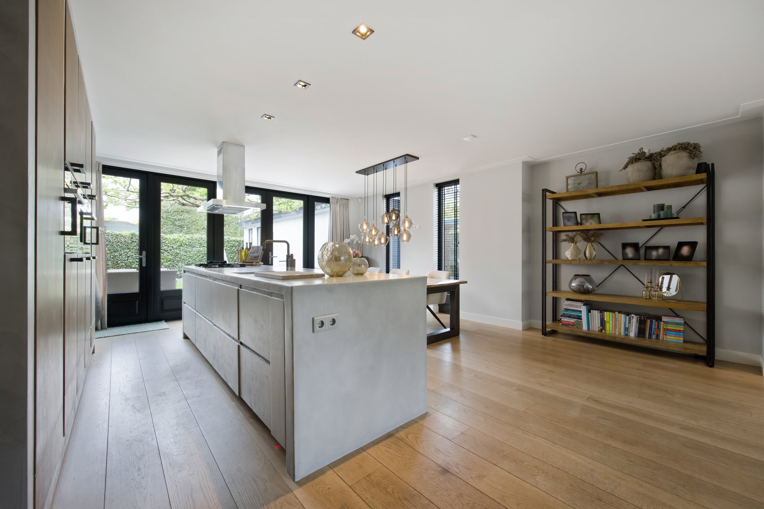 Moderne open keuken en eetkamer met grote ramen, boekenplank, houten vloer en decoraties.