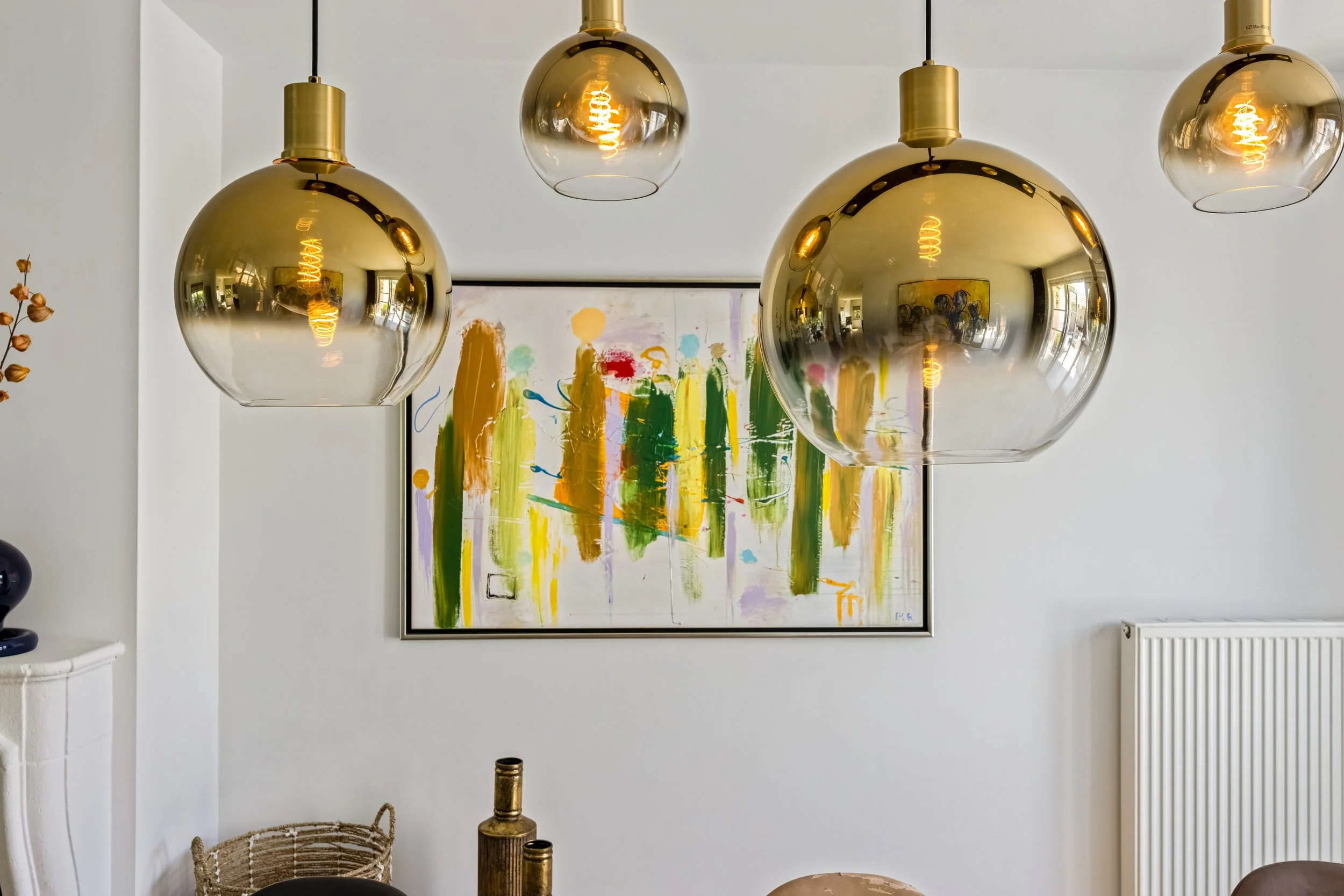 Moderne interieur met vier gouden hanglampen, abstract kunstwerk met felle kleuren op de muur, en decoratieve objecten op tafel.