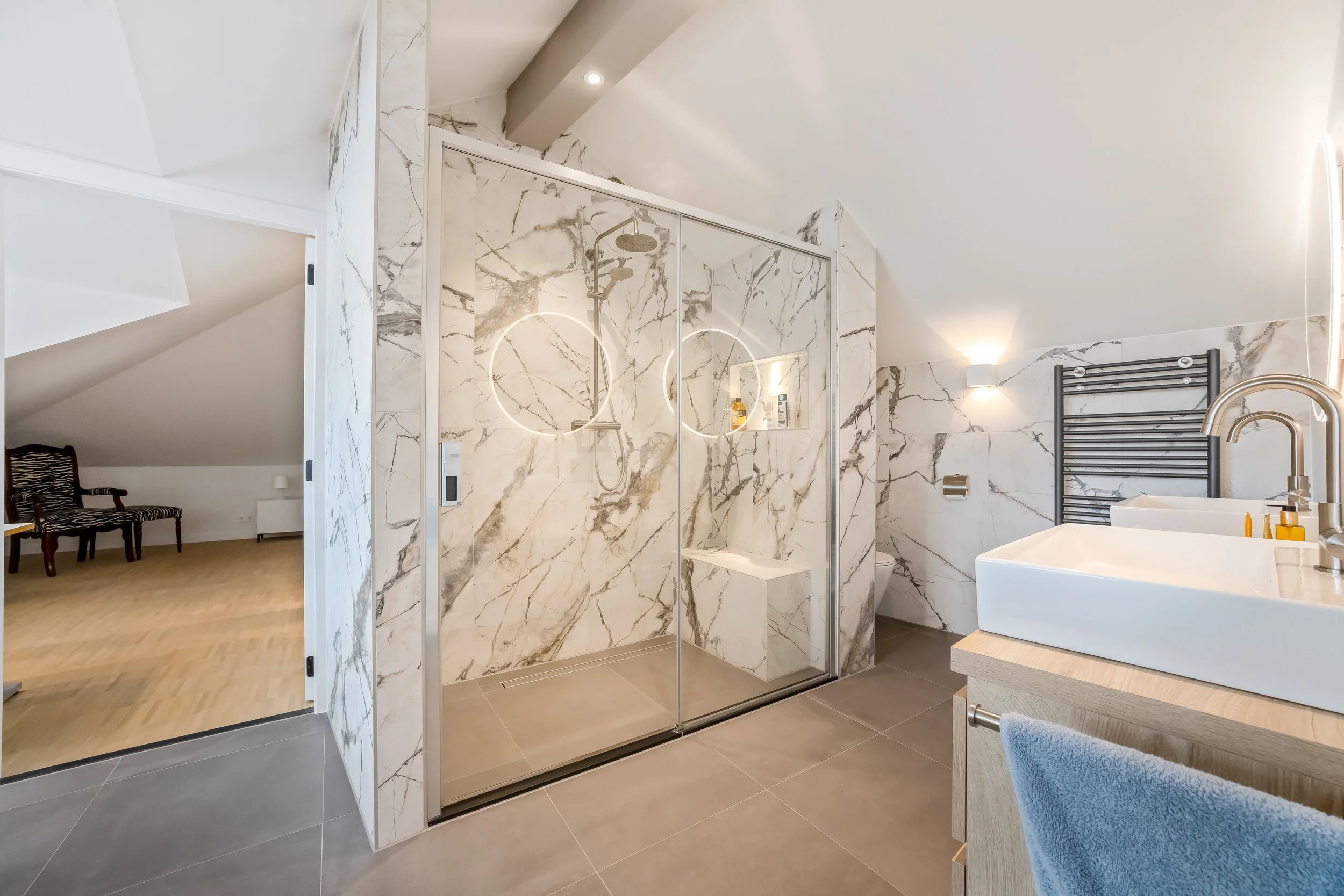 Een moderne badkamer met een glazen inloopdouche met marmeren muren, een wasbak op een houten meubel, een zwart verwarmingsraam, en een toilet. Het plafond is schuin en wit.