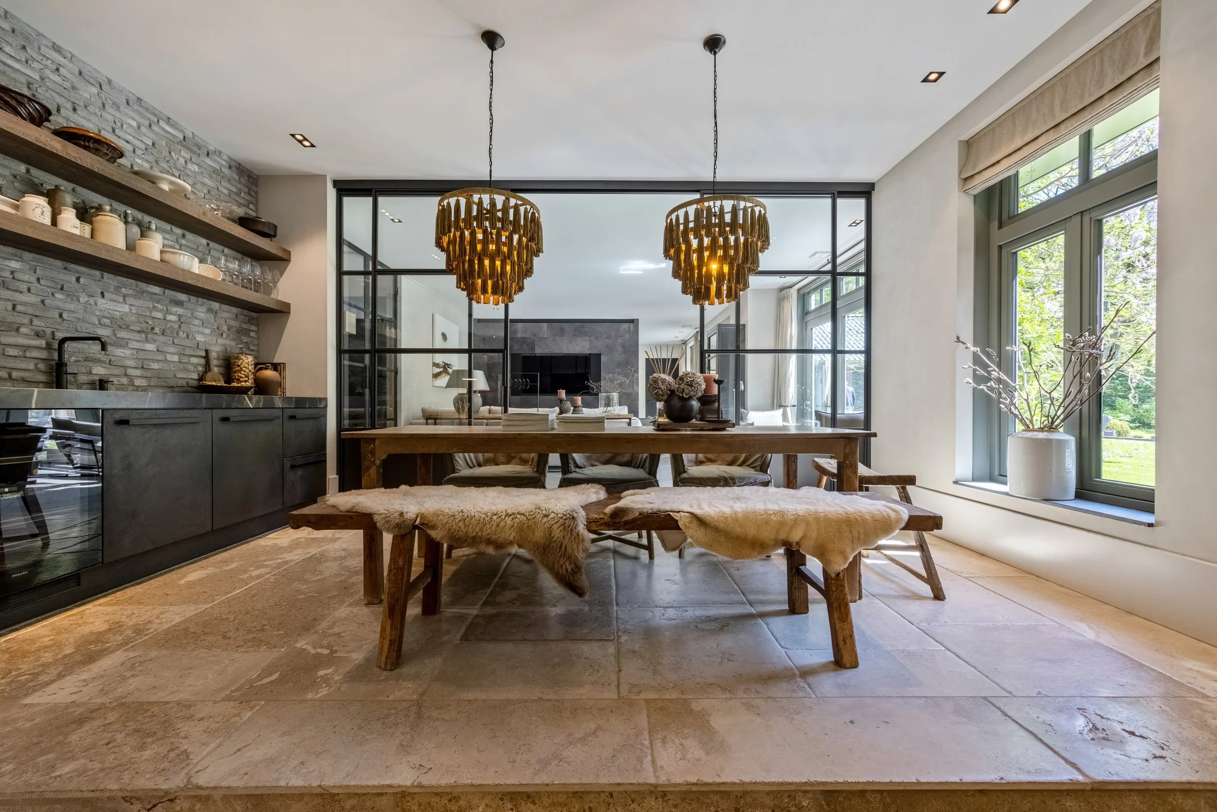 Een modern eetkamertafel met vier houten stoelen en een bank met schapenvacht. Boven de tafel hangen twee gouden hanglampen. Achter de tafel is een grote glazen wand die de eetkamer afscheidt van de woonkamer. Aan de rechterkant is een grote raamkoep
