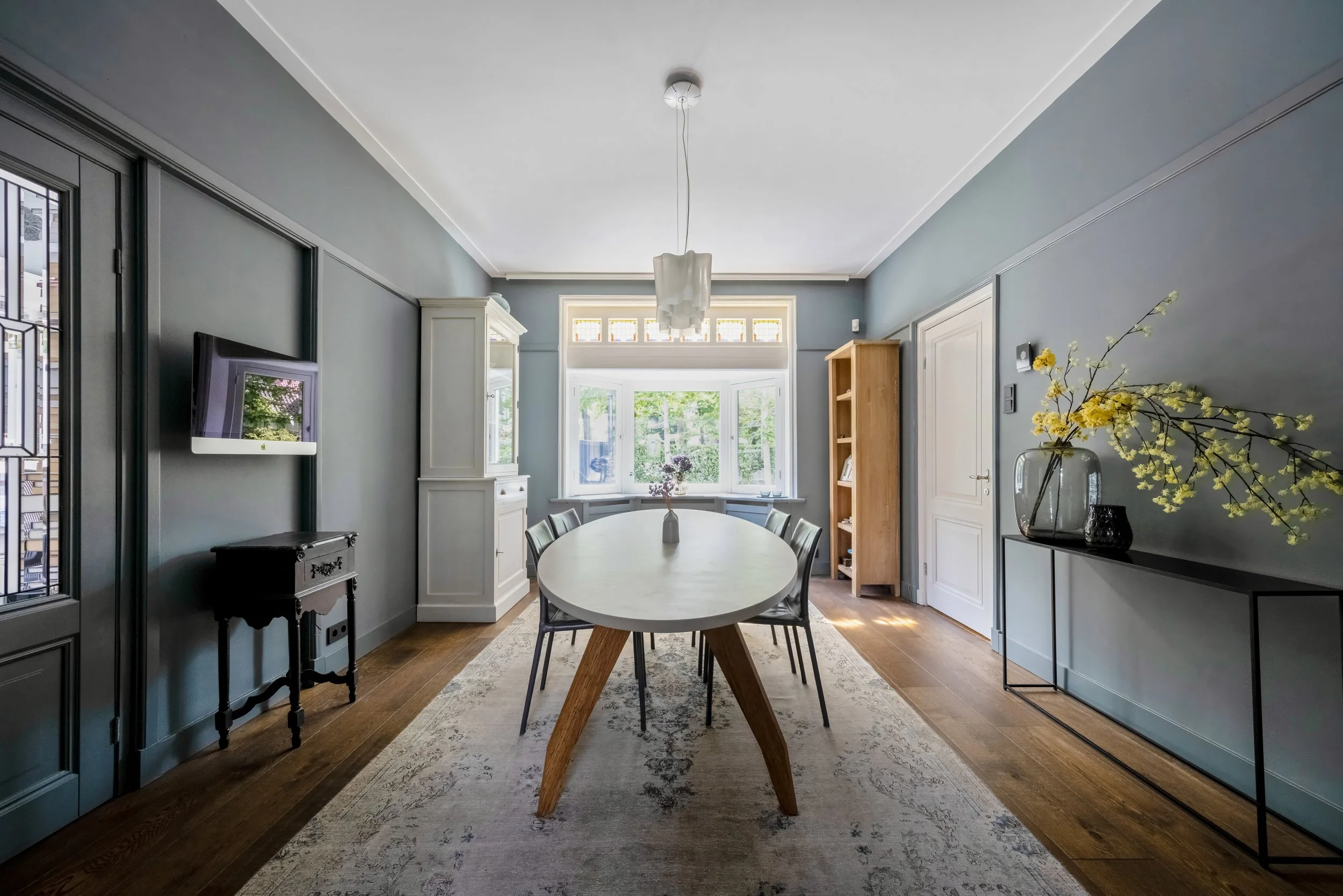 Eethoek in een lichte woonkamer met grote ramen, ronde witte tafel, acht stoelen, een wit buffet, een houten boekenkast, en vazen met bloemen.
