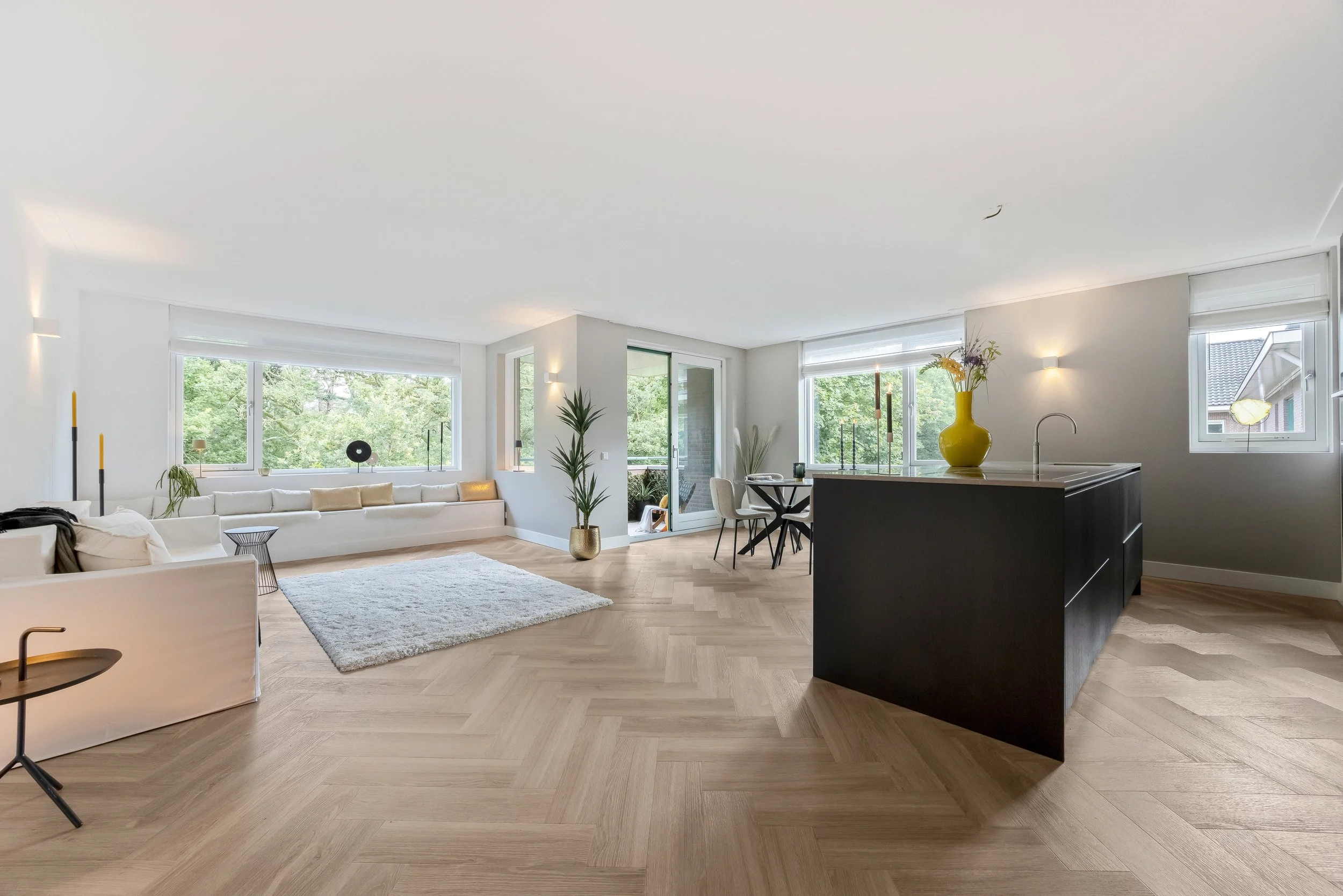 Licht, modern appartement met grote ramen, houten vloer, witte en grijze muren, groene planten, zwart keukeneiland, zithoek met witte bank en een ronde tafel.