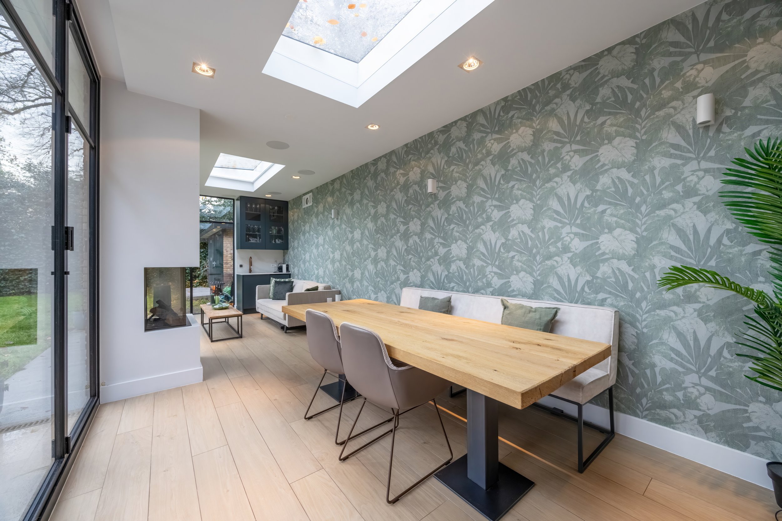 Modern woonruimte met grote glazen schuifdeuren, een houten eettafel, comfortabele banken, een groene plant, muurdecor met een botanisch patroon, en een plafond met daglicht.