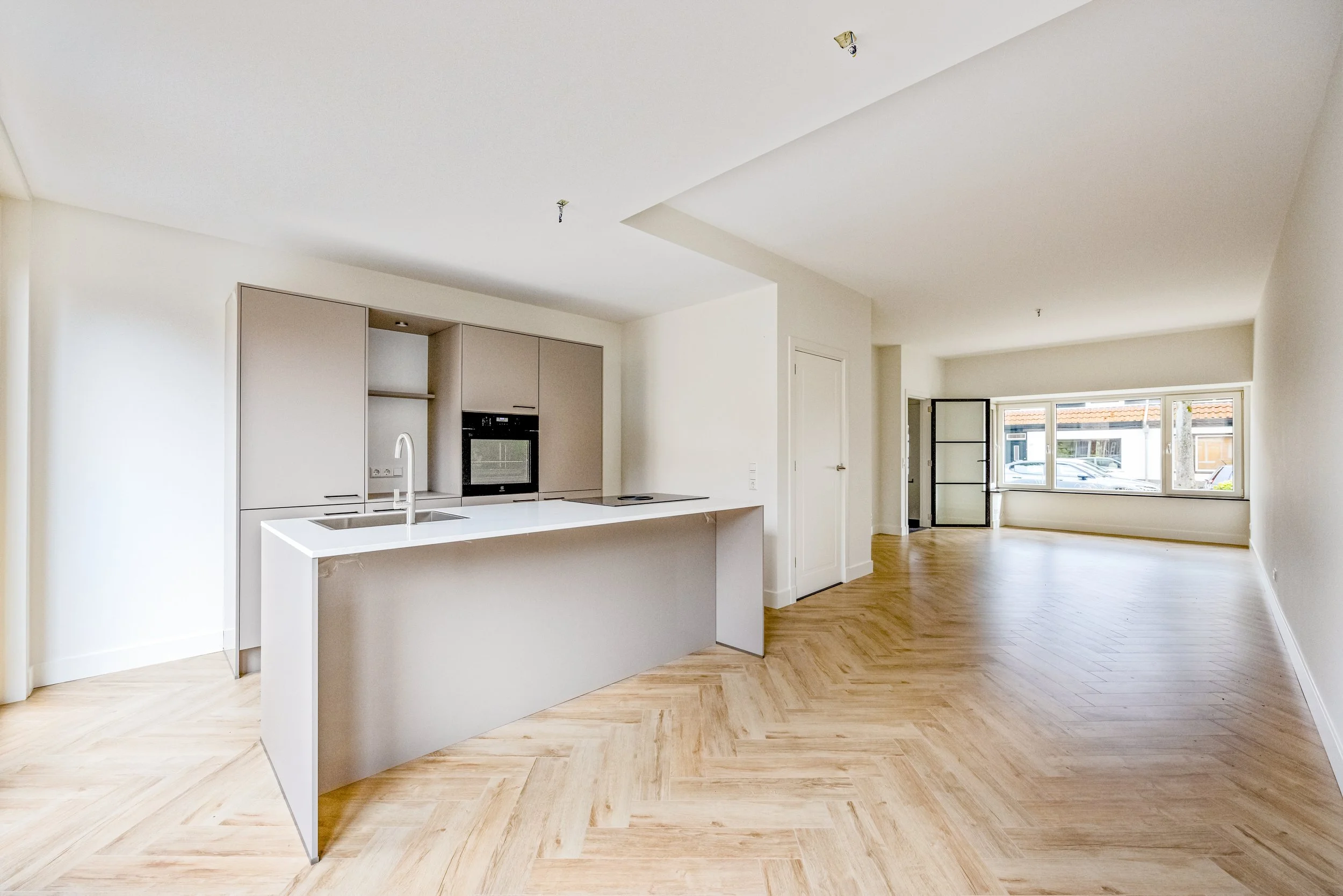 Leeg appartement met open keuken en grote raampartij, houten vloer en witte muren.
