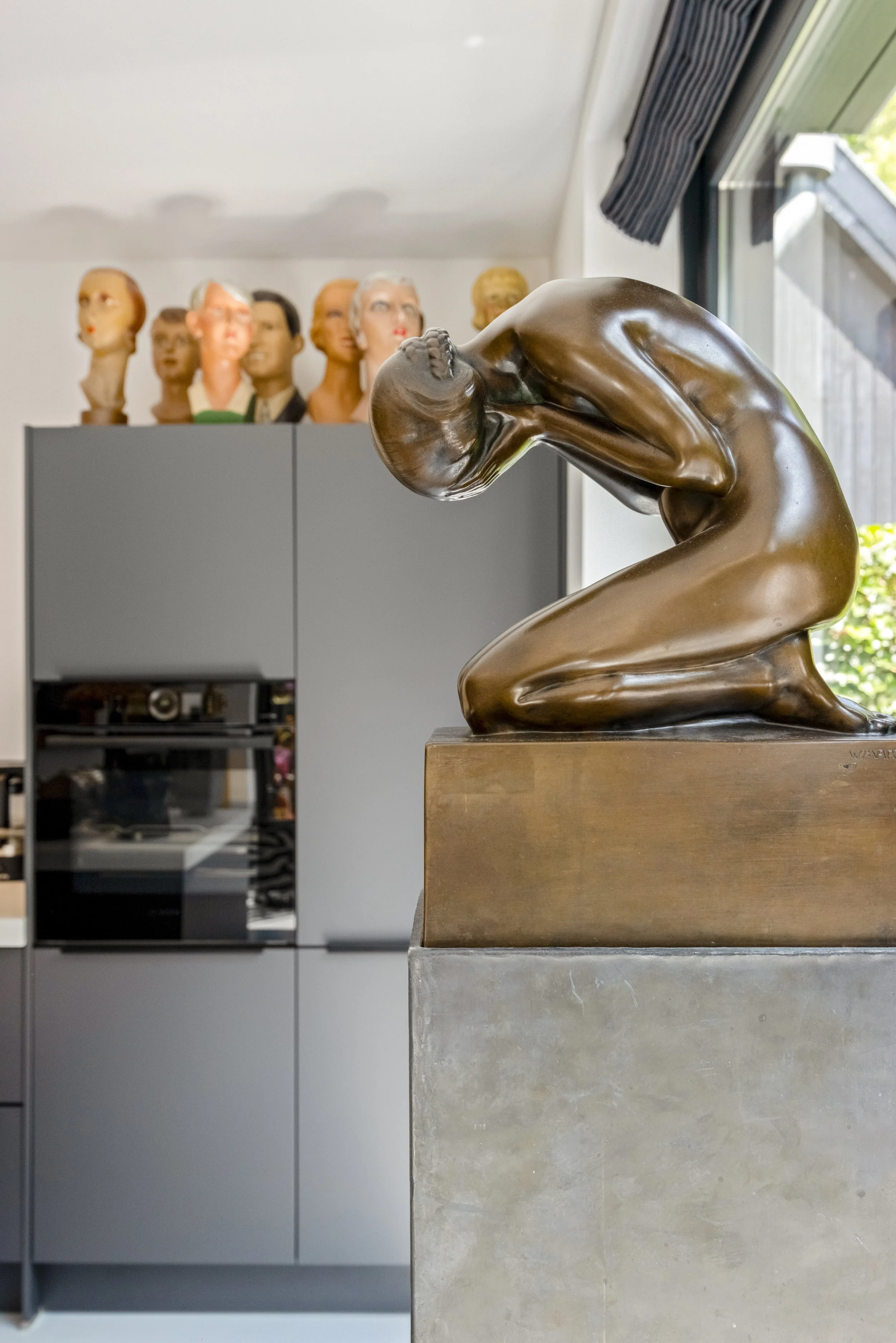 Een bronzen beeld van een vrouw die knielt en haar hoofd en handen over haar oren houdt, geplaatst op een stenen sokkel. Op de achtergrond staan verschillende poppenhoofden en een keukenkastje.