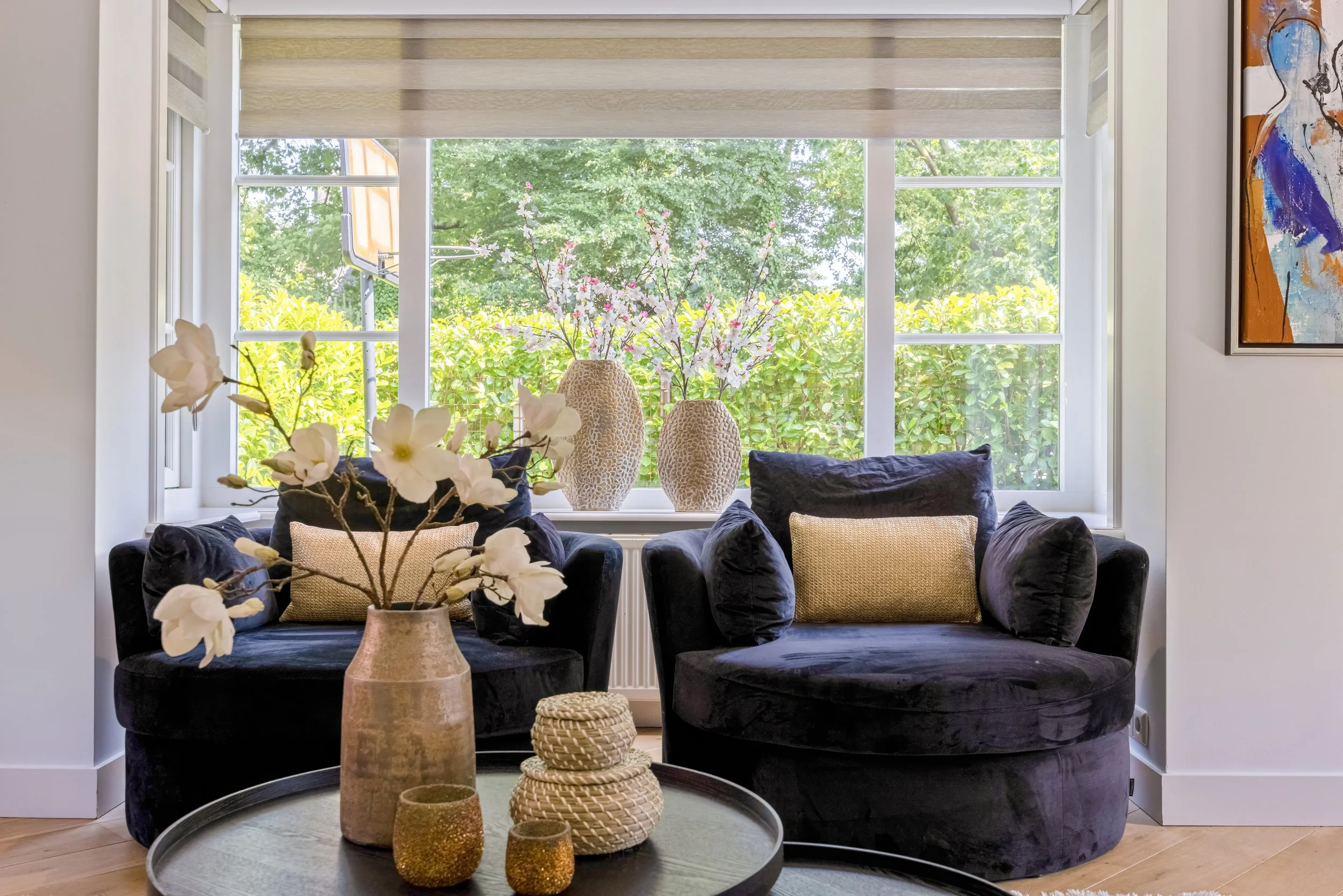 Woonkamer met twee zwarte fauteuils, vazen met bloemen op de vensterbank, een tafel met decoratieve voorwerpen, en een groot raam met uitzicht op groene bomen en struiken.