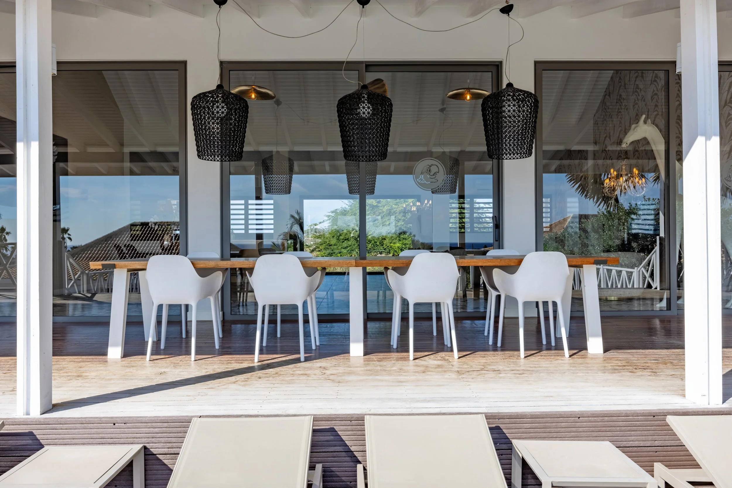 Een moderne terrasruimte met een lange houten tafel en witte stoelen, grote zwart opengewerkte hanglampen, en een glazen muur met uitzicht op een tuin en het water.