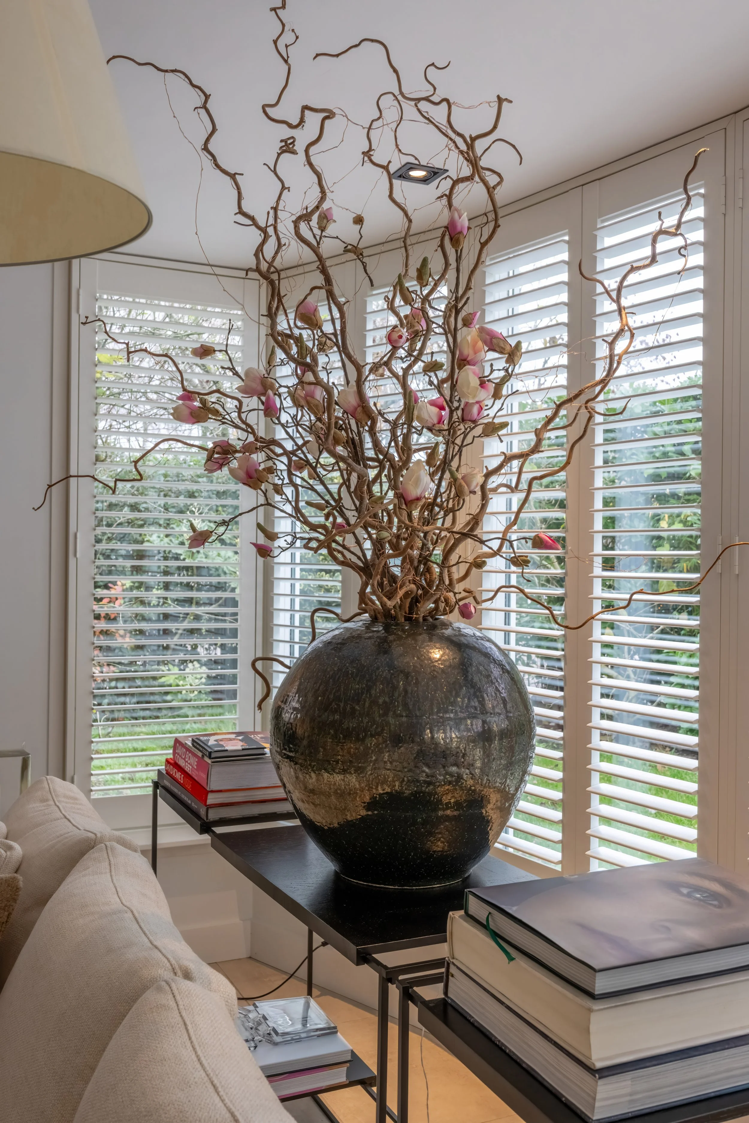 Een grote vaas met gedraaide takken en roze bloemen, geplaatst op een zwarte tafel in een woonkamer met witte jaloezieën.