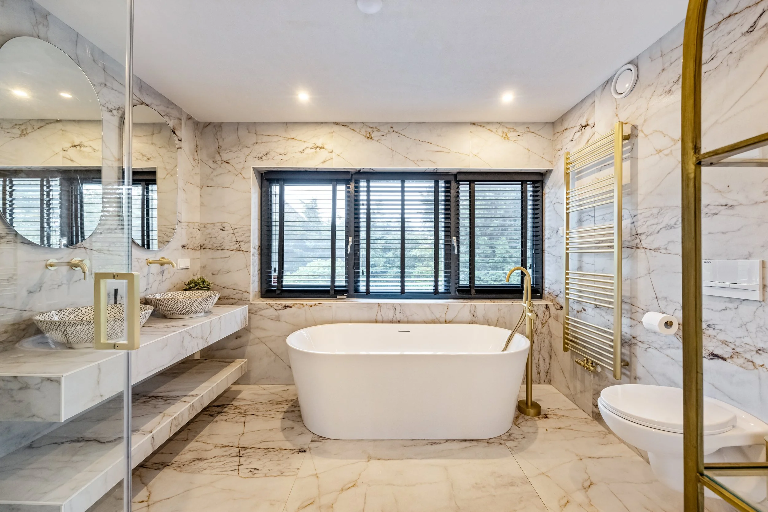Luxe badkamer met marmeren wanden en vloer, vrijstaande badkuip onder grote raam met zwarte jaloezieën, dubbele wastafel met ronde spiegels, radiator en toilet.