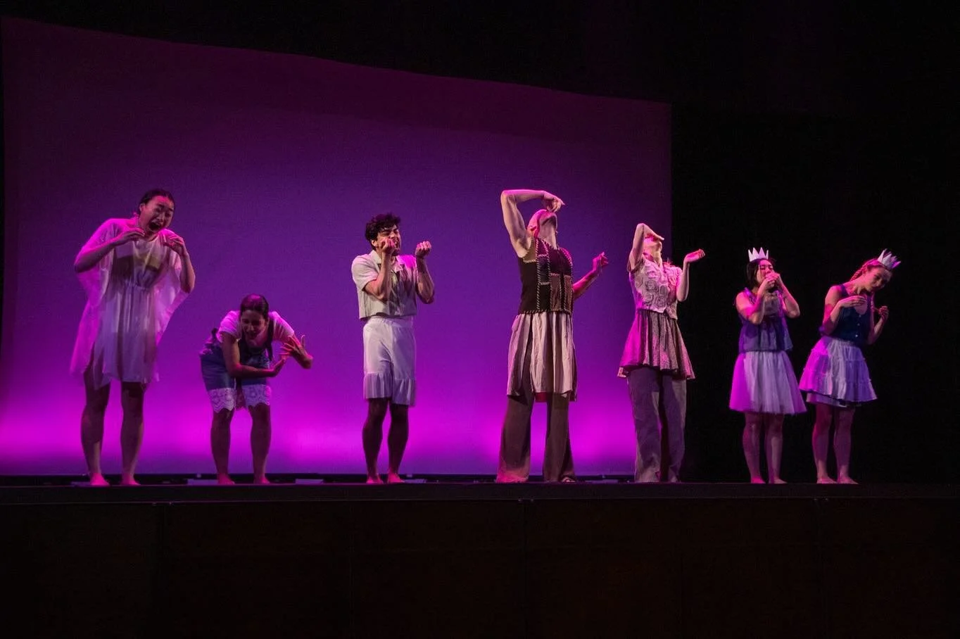Group of seven people on stage performing a play or dance, some wearing paper crowns, with a purple background lighting.