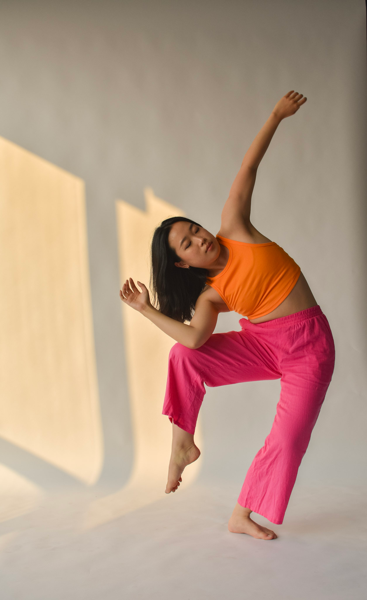 A woman in an orange tank top and pink pants is dancing or performing a movement, with one arm raised above her head and the other bent behind her head. She is balancing on one leg with her other leg lifted. The background is plain with shadow patterns.