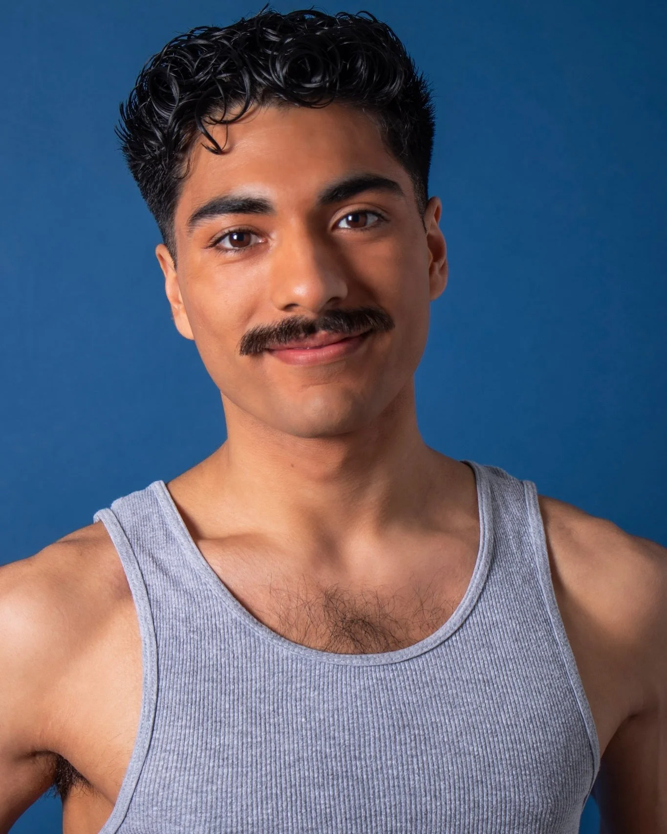 Portrait of a young man with dark, curly hair, a mustache, wearing a grey tank top, standing against a blue background.