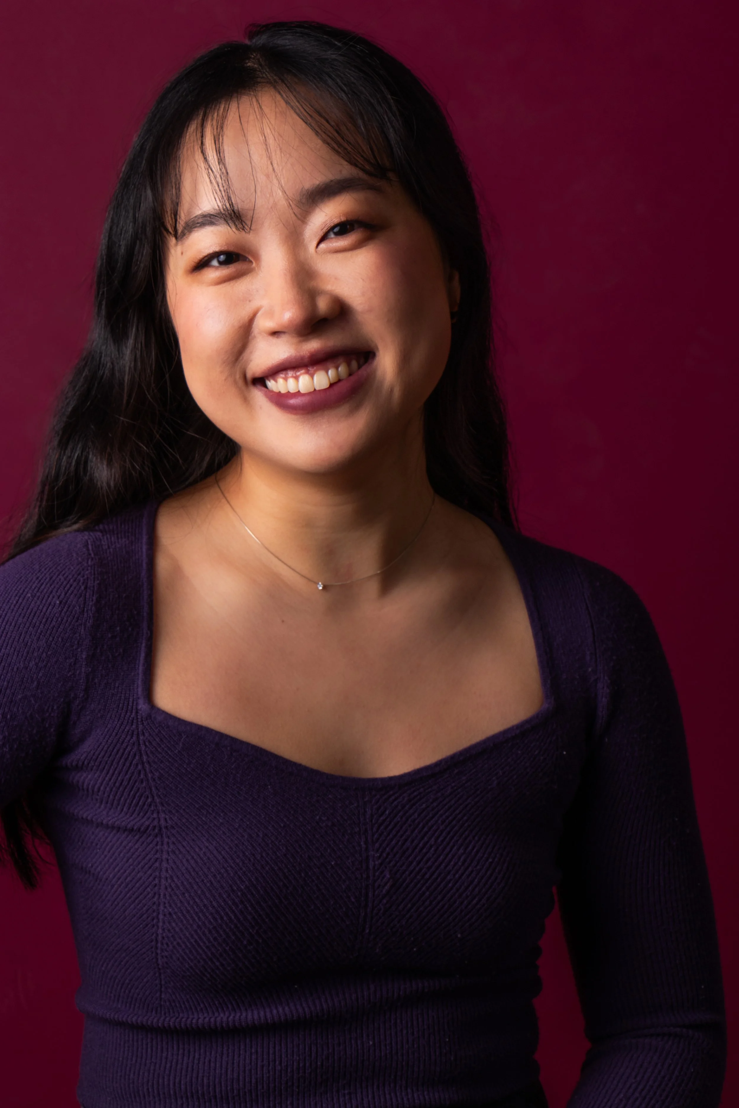 Rachel Ha-Eun Lee wearing a purple top and a delicate necklace, posed against a deep red background.