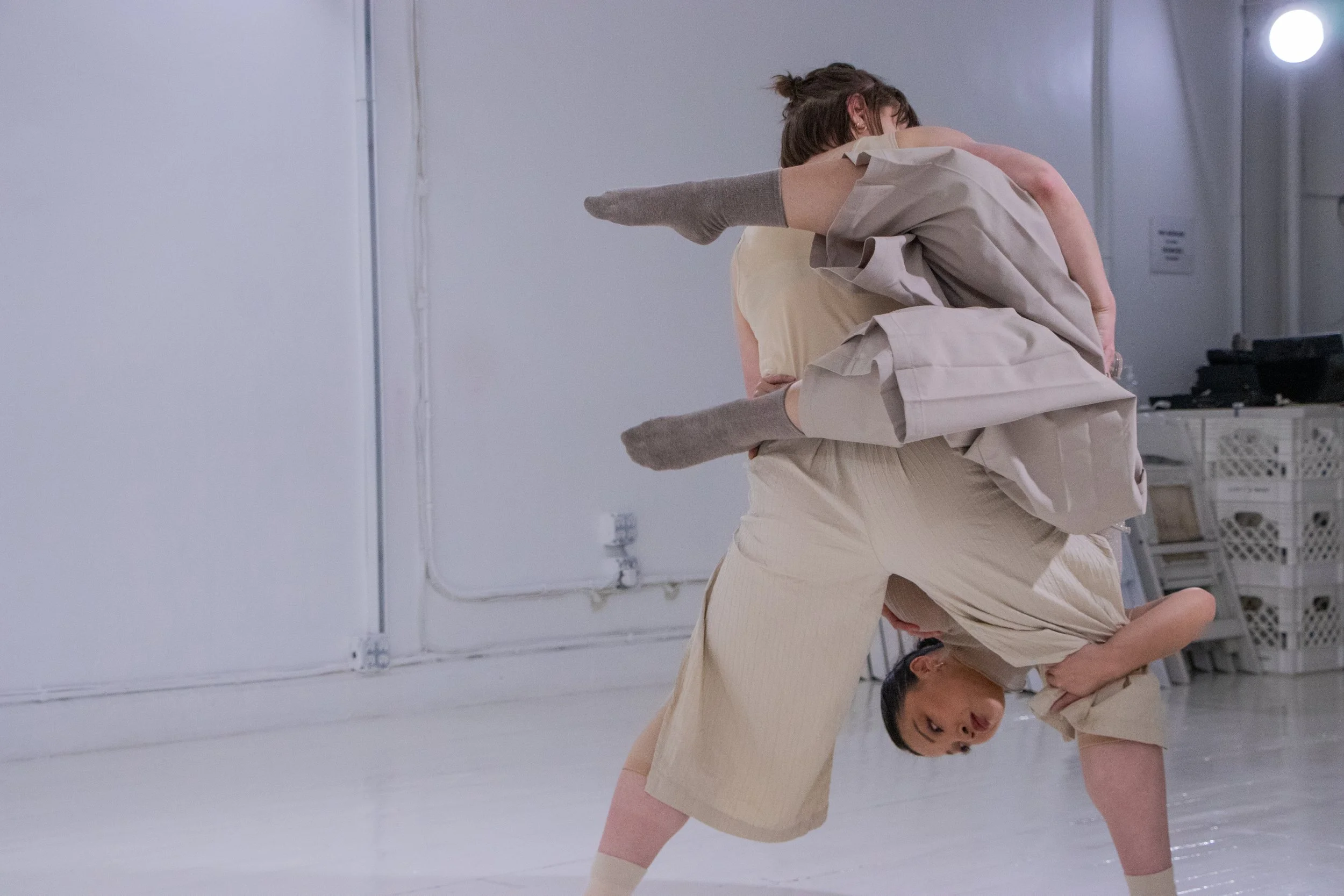 Two women practicing a dance or martial arts move in a minimalistic, white room. One woman bends forward on her hands and one knee, while the other woman is draped over her back, holding her hips.