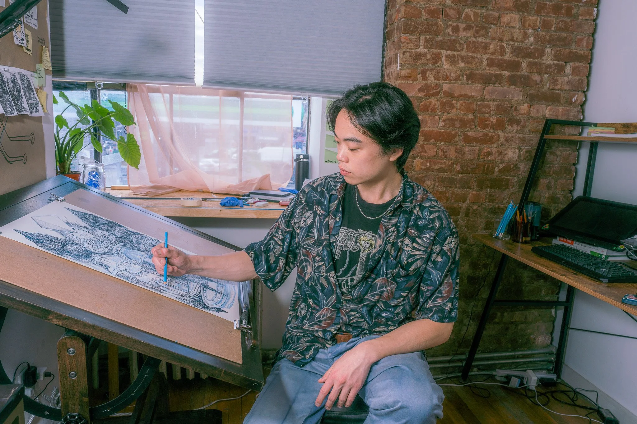 Young man drawing detailed artwork on a tilted drafting table in a studio with exposed brick wall and window.