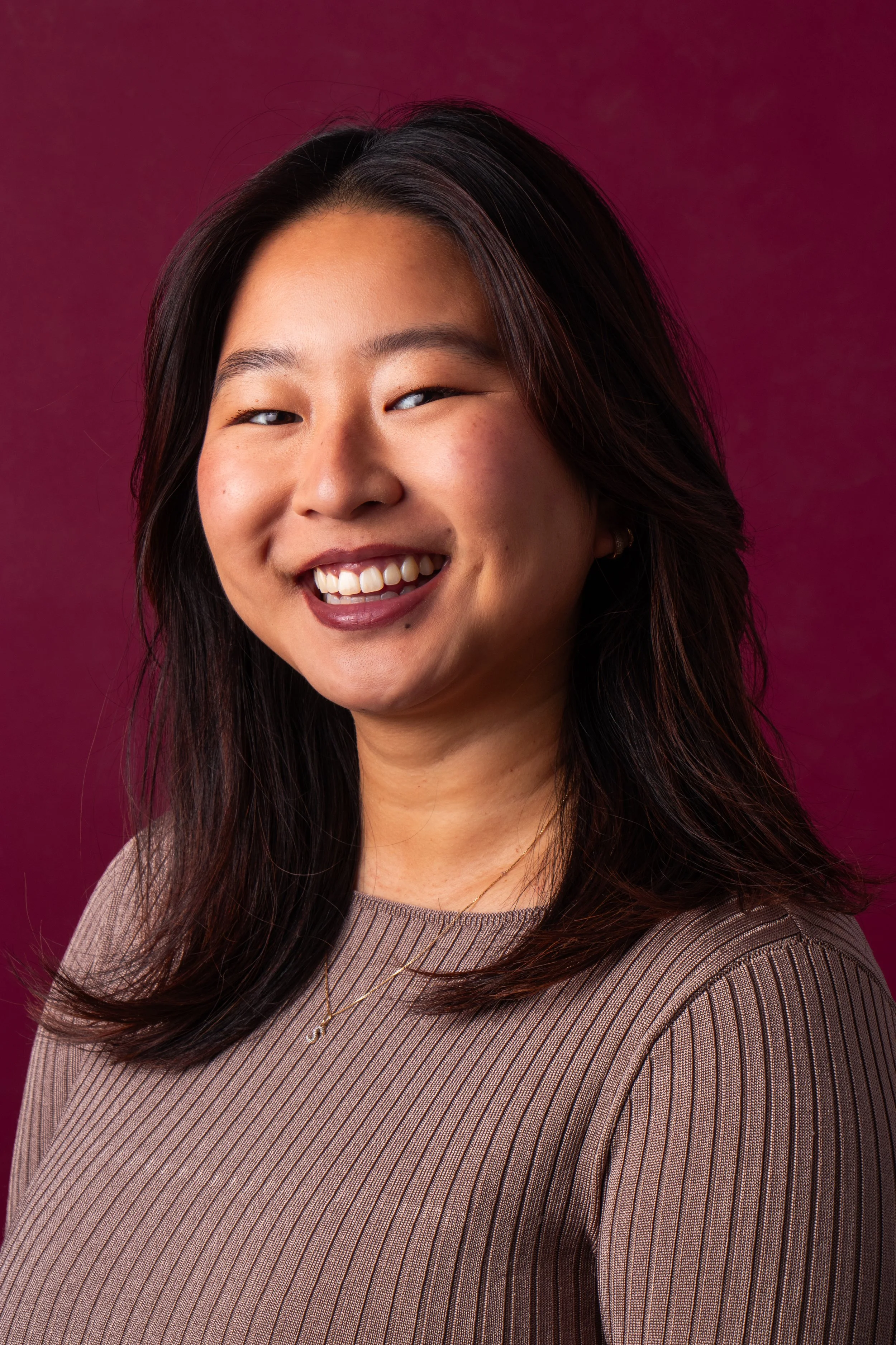 Close-up portrait of a young Asian woman with shoulder-length dark hair, smiling, wearing a beige ribbed top and a gold necklace, against a dark pink background.