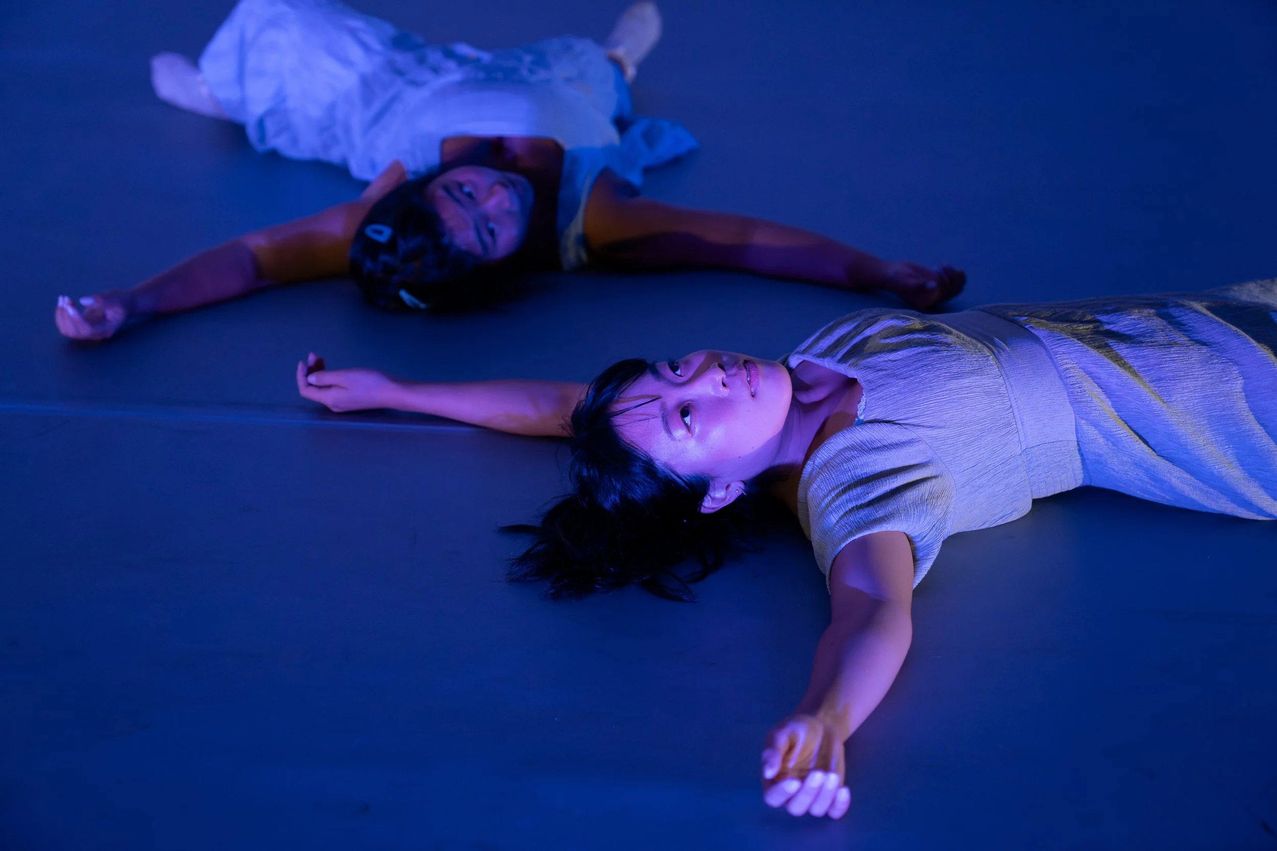 Rachel and Stephanie Shin are lying on a dark stage or floor, illuminated by purple and blue lighting, with their arms extended outwards.
