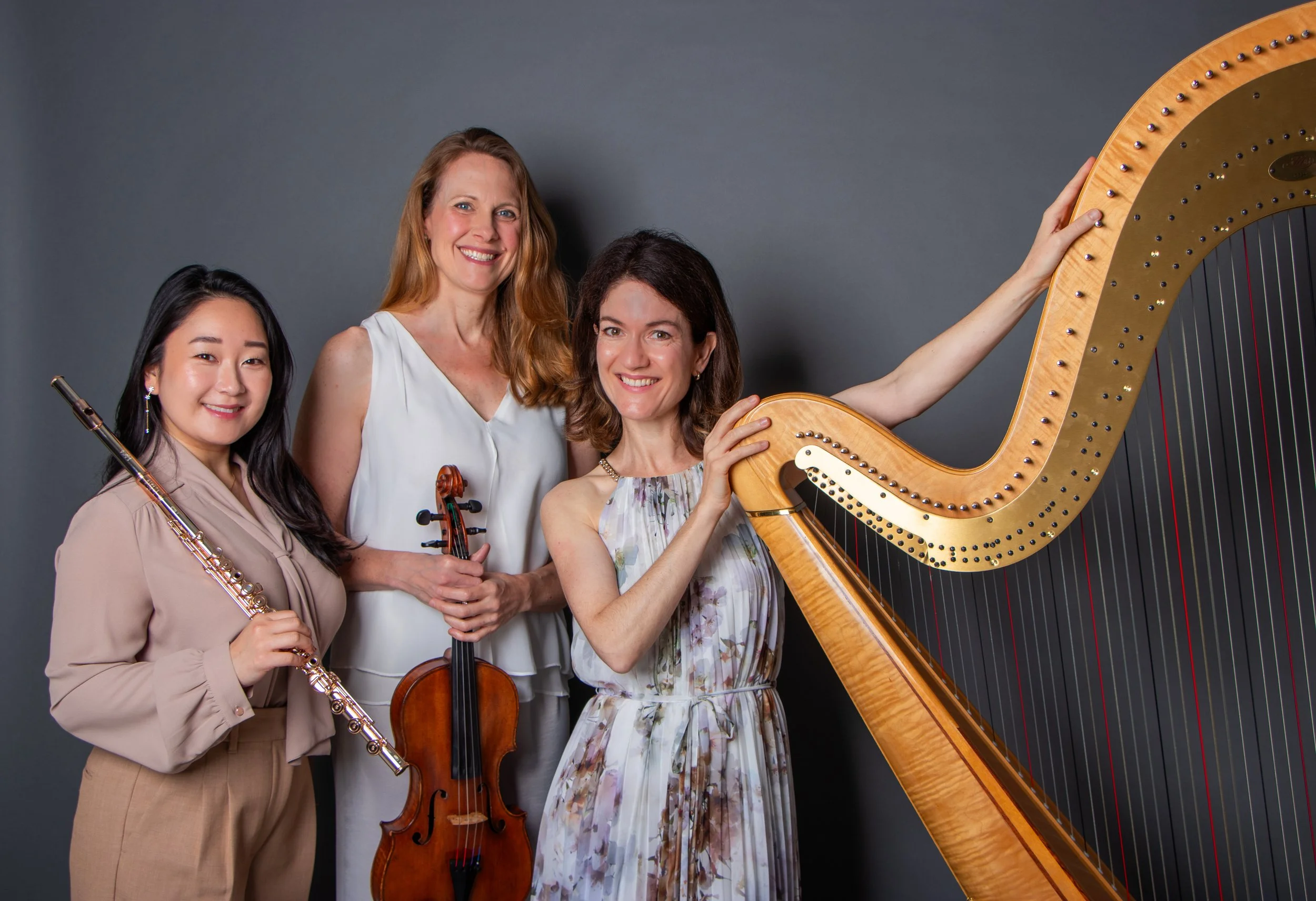 Group of four women musicians posing with their instruments against a dark gray background, including a flute, violin, and harp.