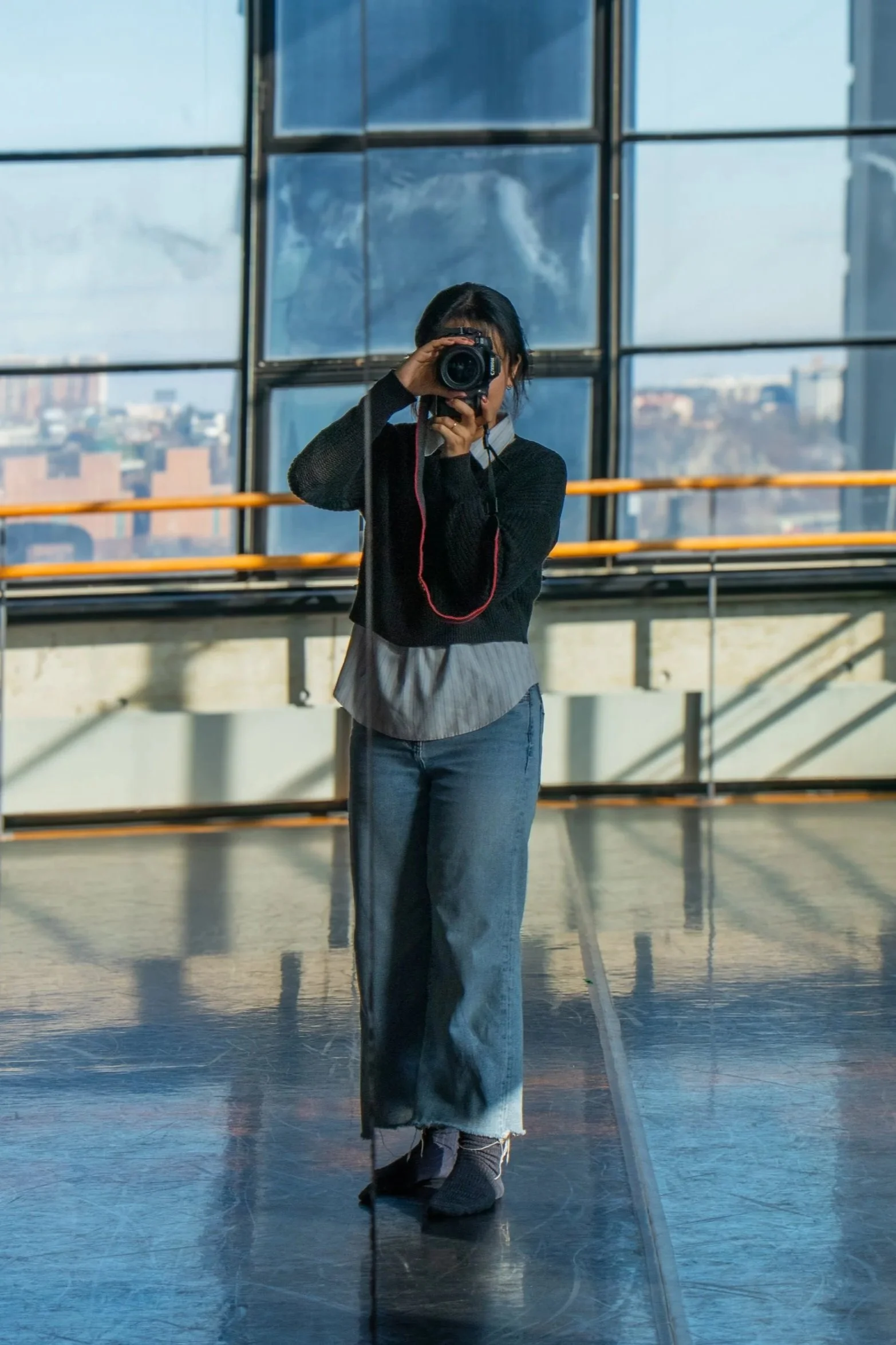 Person taking a photograph in front of a large window with cityscape view, wearing a black sweater, gray shirt, and jeans.