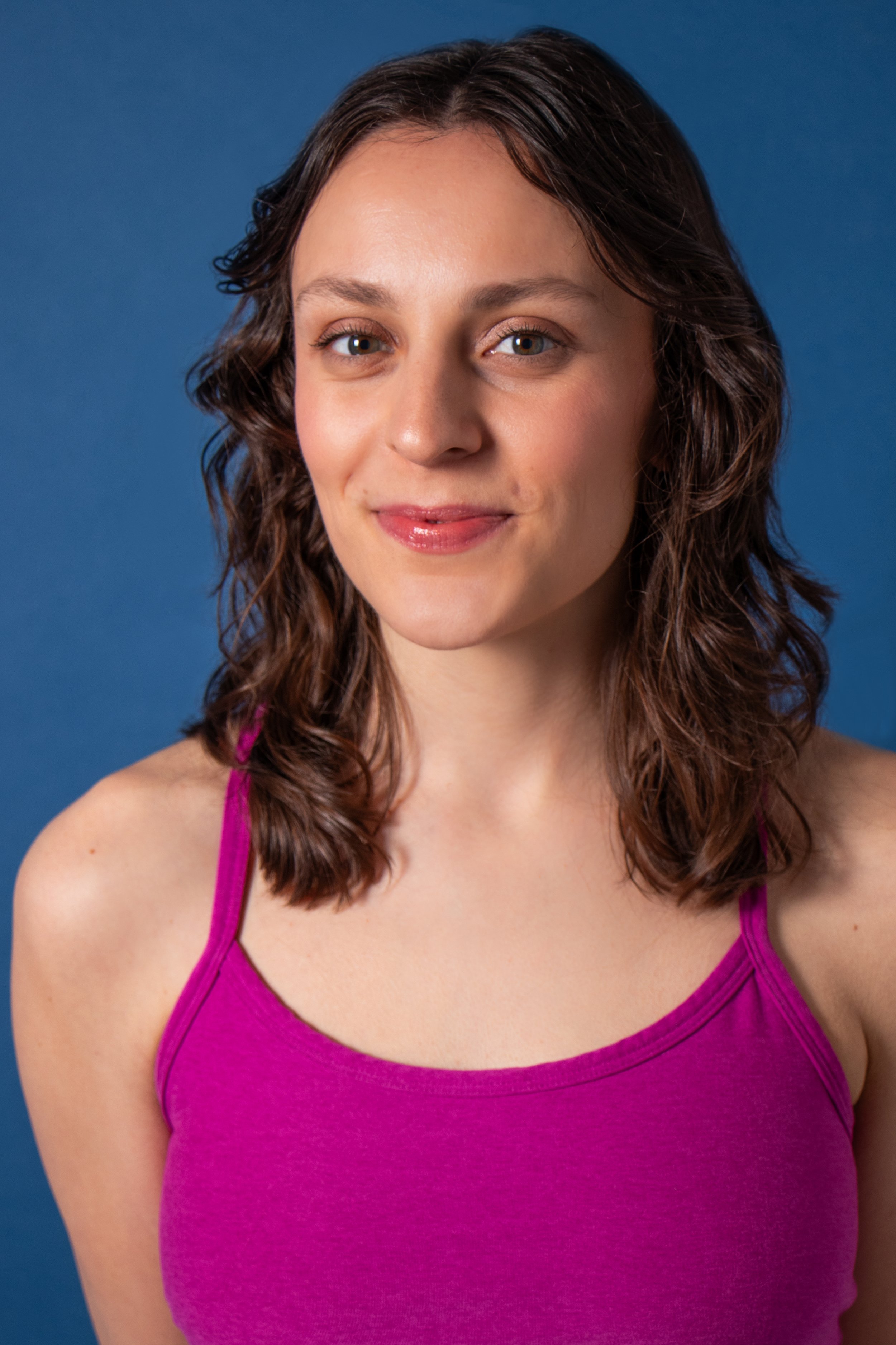 A young woman with shoulder-length curly brown hair, wearing a bright pink tank top, smiling softly against a solid blue background.