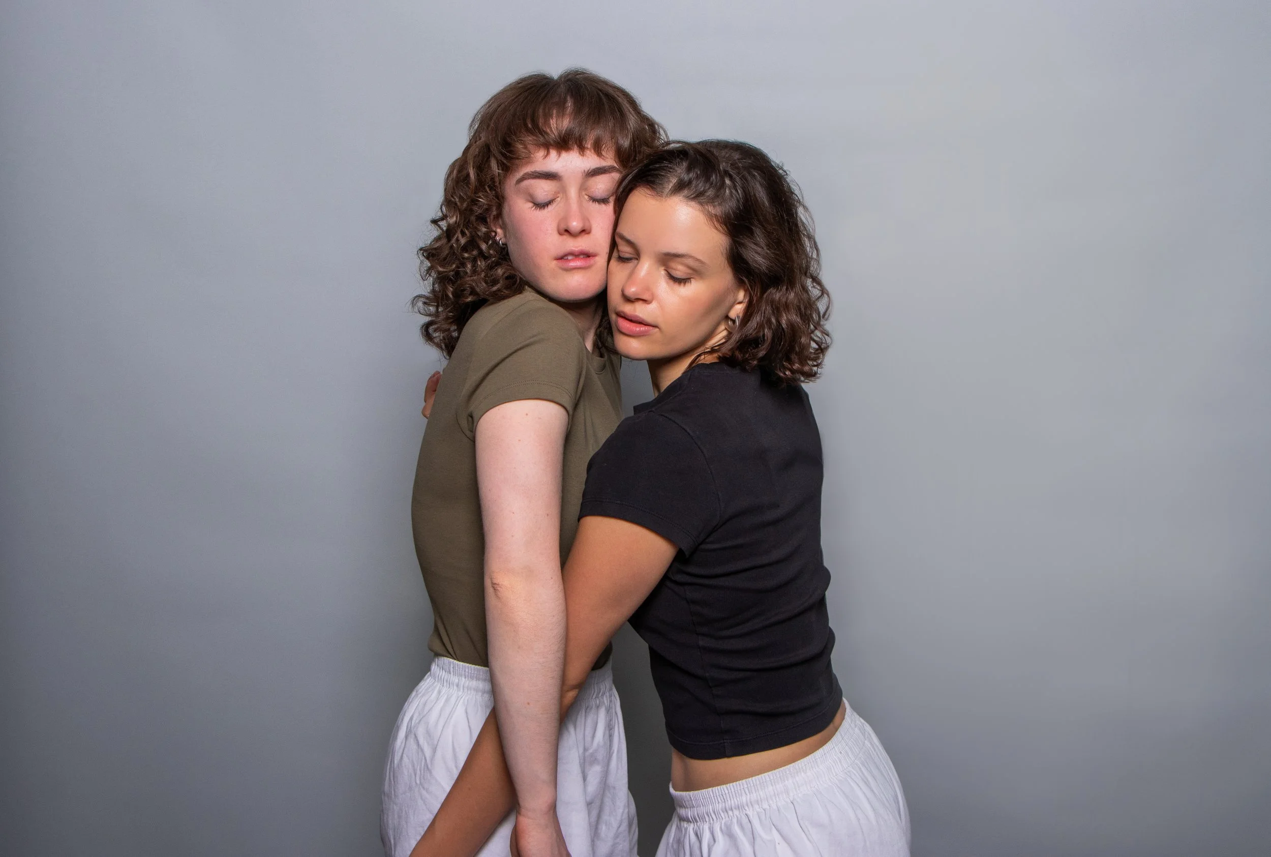 Two women with curly hair hugging each other against a plain, neutral background.