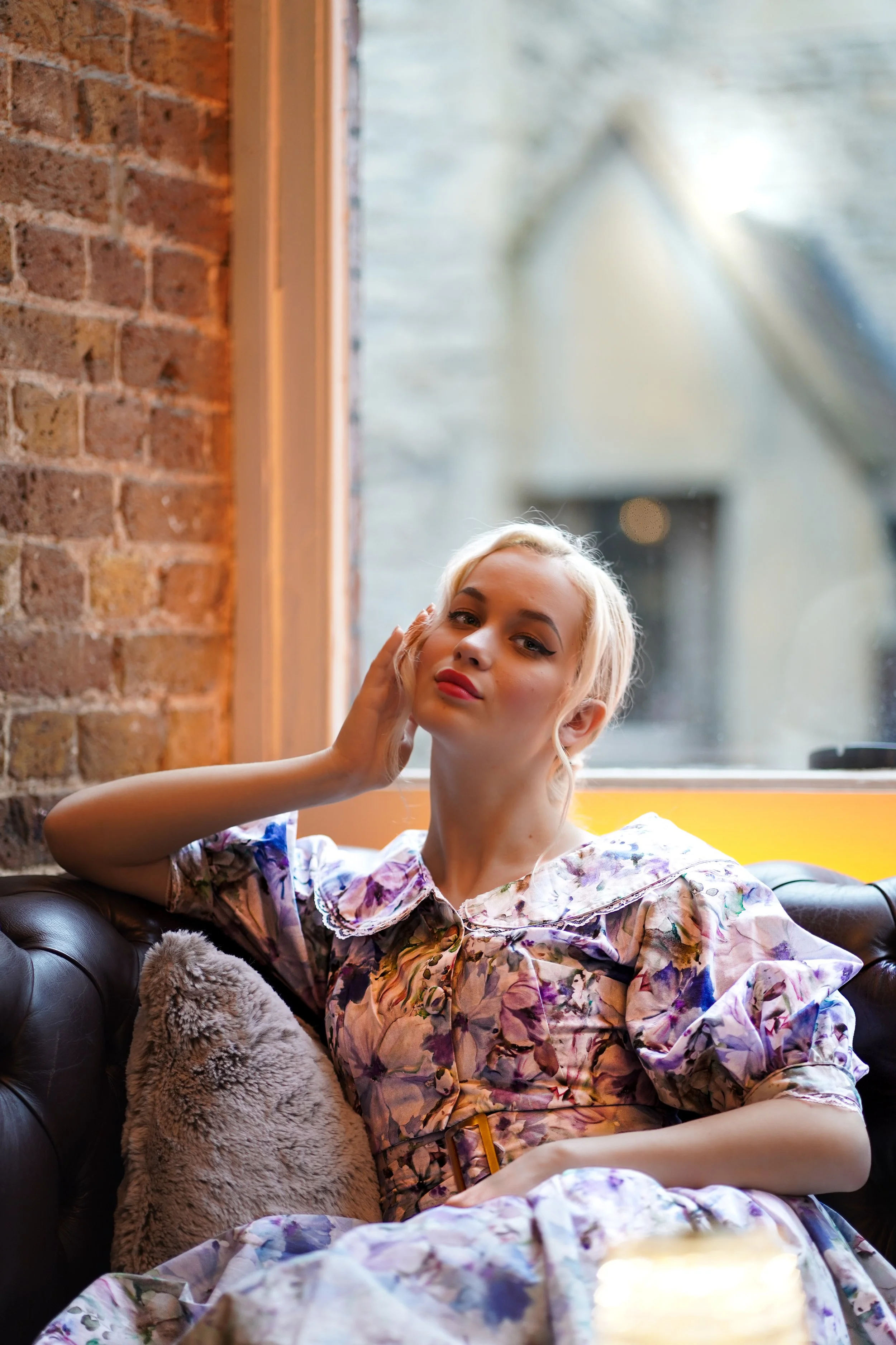 A young woman with platinum blonde hair and makeup, wearing a floral dress, relaxing on a leather sofa by a window in a cozy cafe with brick walls.